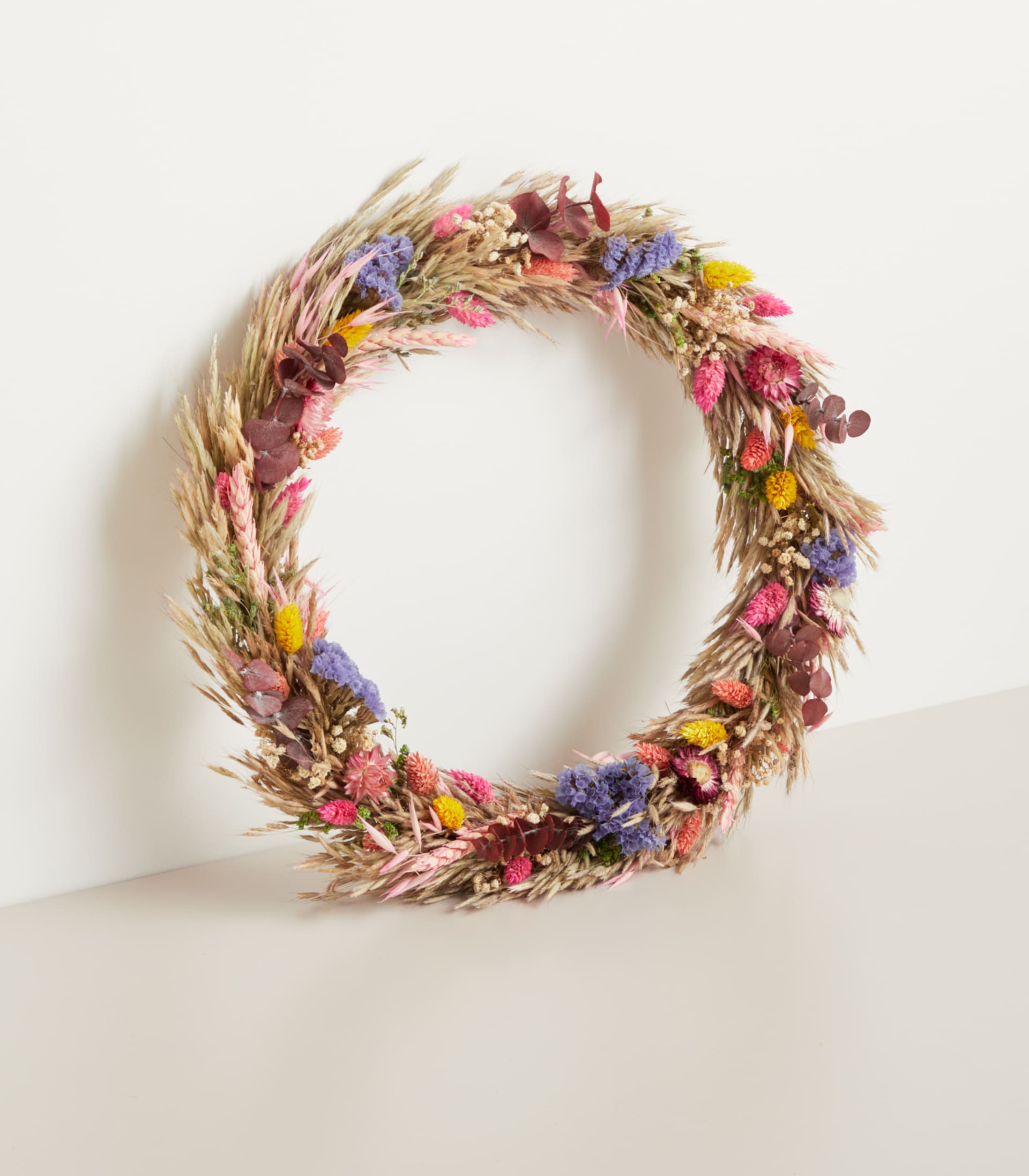 Ein lebendiger, runder Kranz aus getrockneten Blumen, Gräsern und anderen natürlichen Elementen wird vor einem schlichten, hellen Hintergrund präsentiert.