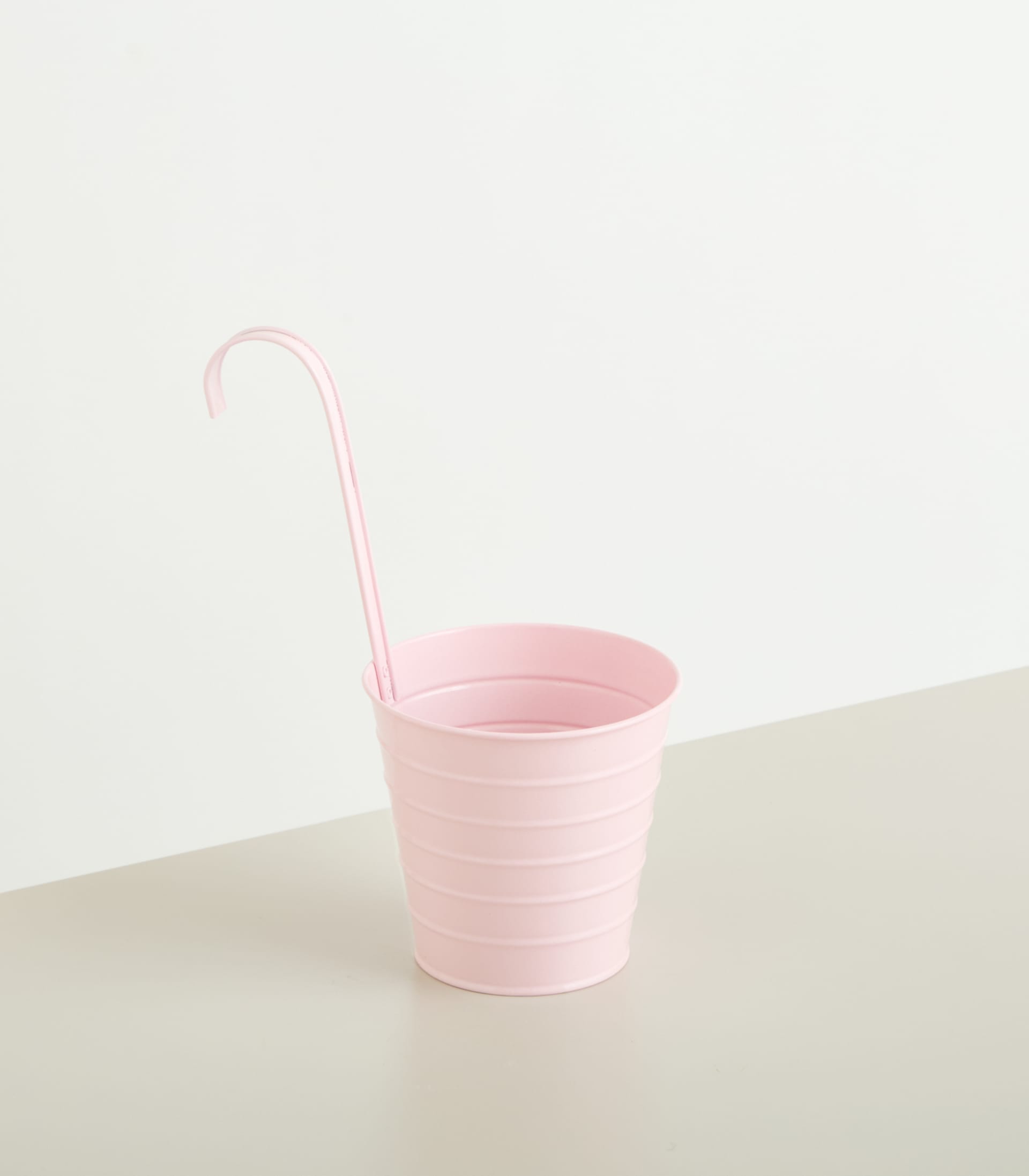 Eine rosafarbene Gießkanne aus Metall mit gebogenem Griff steht auf einer hellen Oberfläche vor einem schlichten weißen Hintergrund.