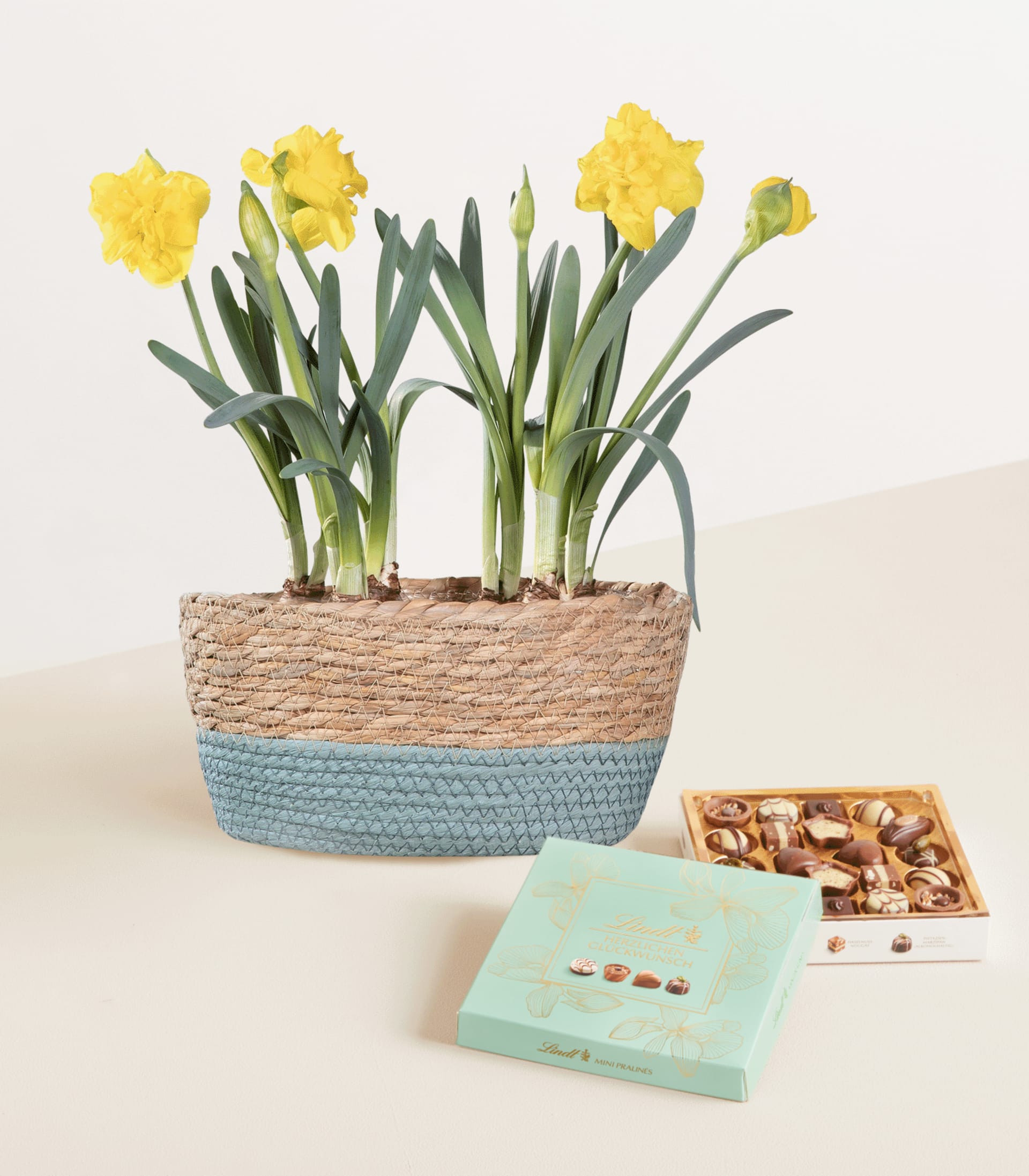 Auf einer hellen Oberfläche steht ein Korb mit leuchtend gelben Narzissen, daneben eine kleine Schachtel Pralinen und ein dekoratives Tablett.