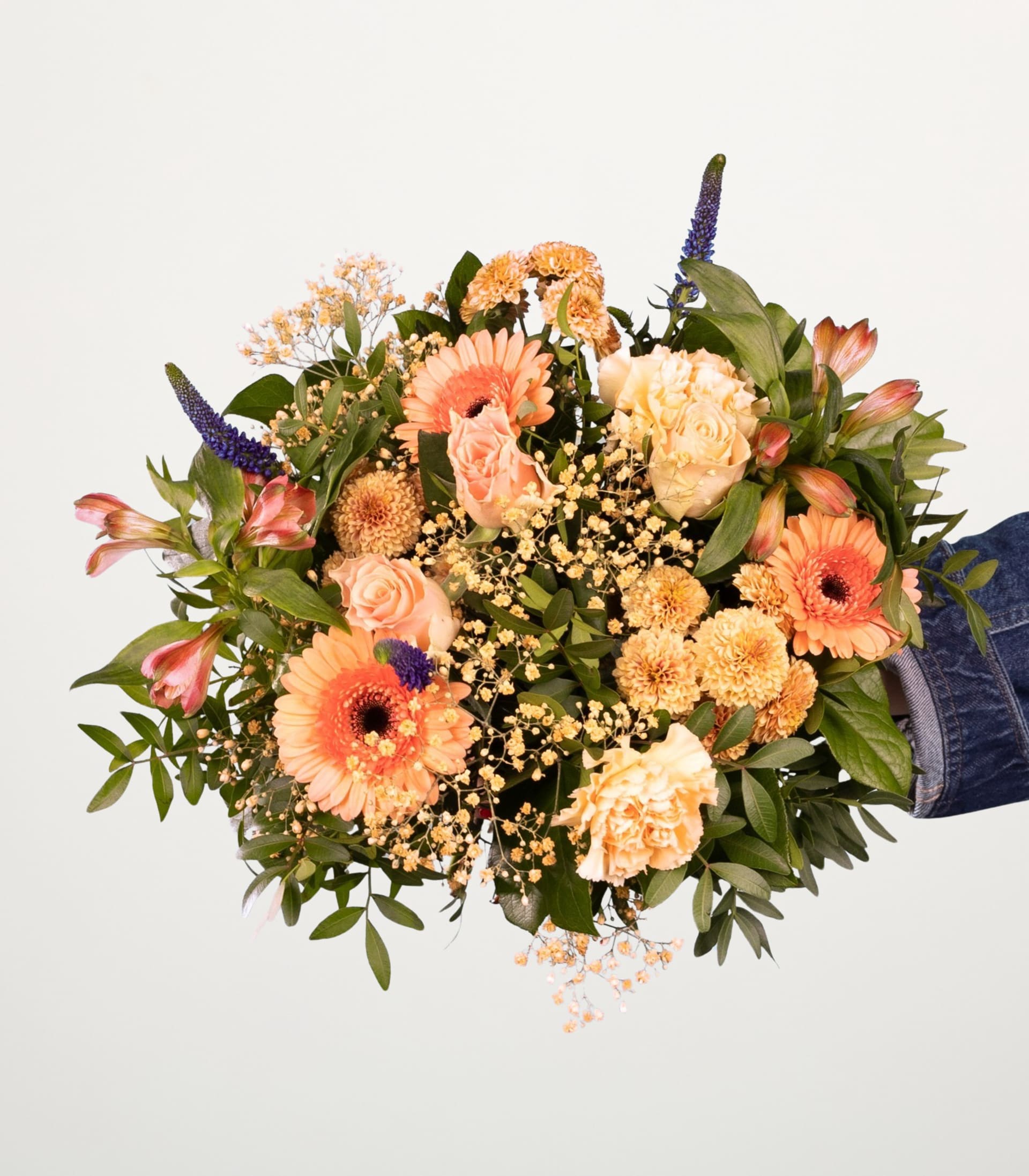 Ein farbenfroher Strauß verschiedener Blumen, darunter Gerbera, Rosen und Nelken, wird vor einem schlichten weißen Hintergrund in den Händen einer Person gehalten.