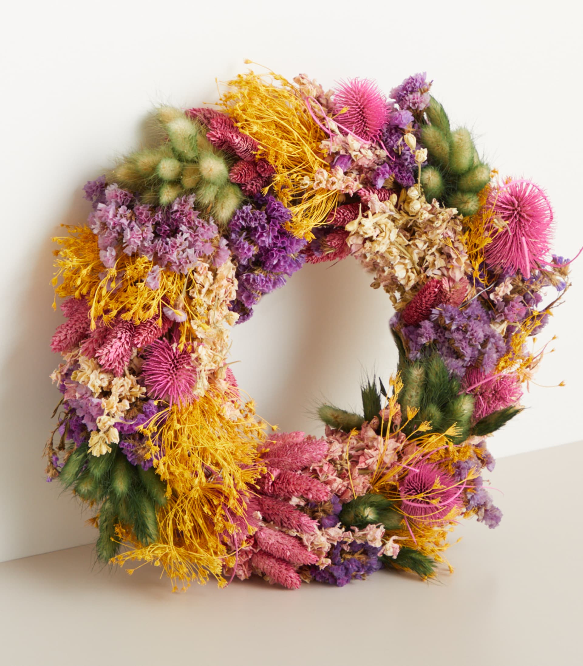 Ein leuchtender, runder Kranz aus verschiedenen getrockneten und frischen Blumen, darunter in Gelb-, Rosa- und Lilatönen, liegt auf einer schlichten, hellen Oberfläche.