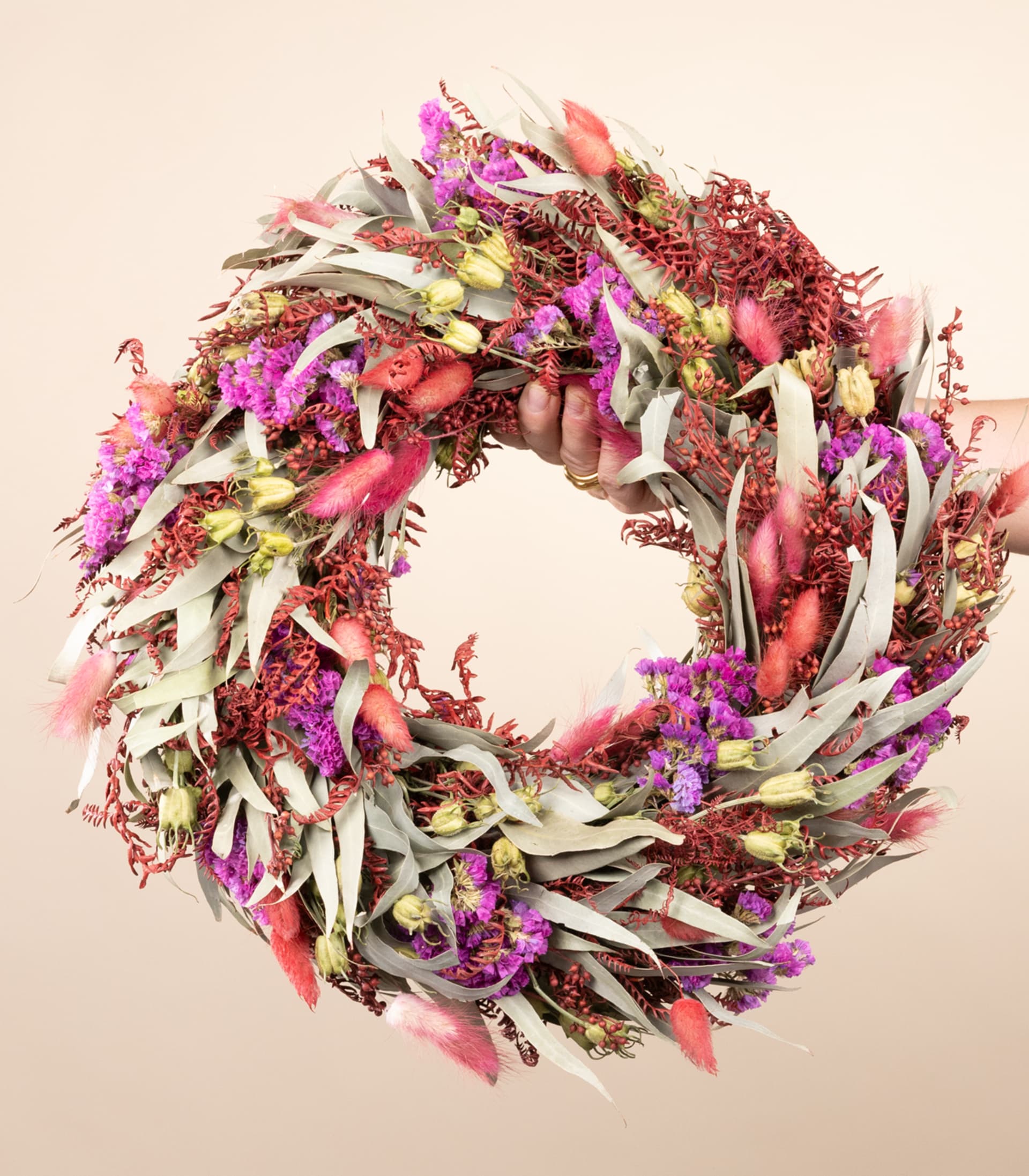 Ein lebendiger, farbenfroher Kranz aus verschiedenen getrockneten Blumen und Blättern, der vor einem neutralen beigen Hintergrund in den Händen einer Person gehalten wird.