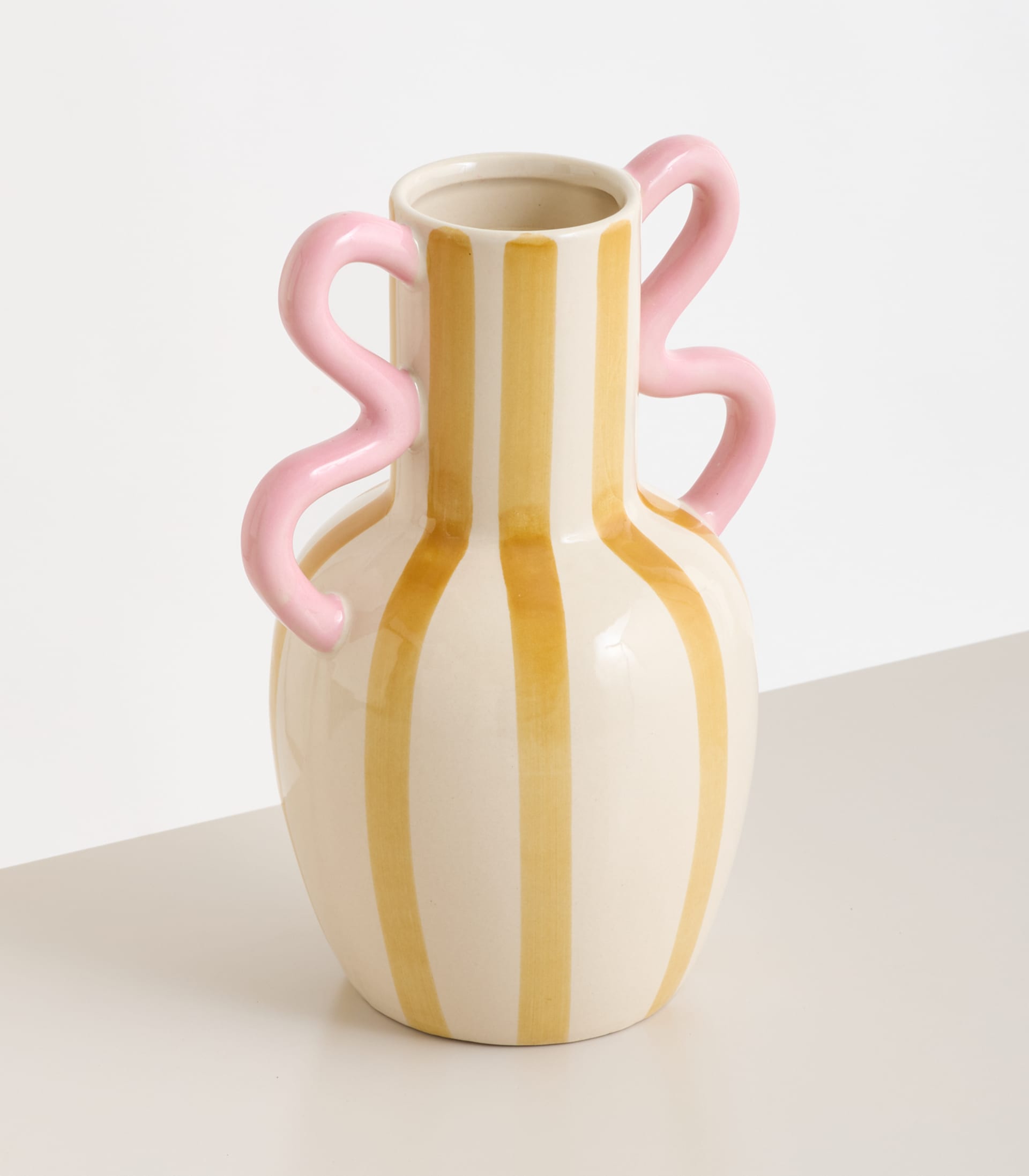 Eine gelb-weiß gestreifte Keramikvase mit rosa Henkeln steht auf einer neutralfarbenen Fläche vor einem schlichten Hintergrund.