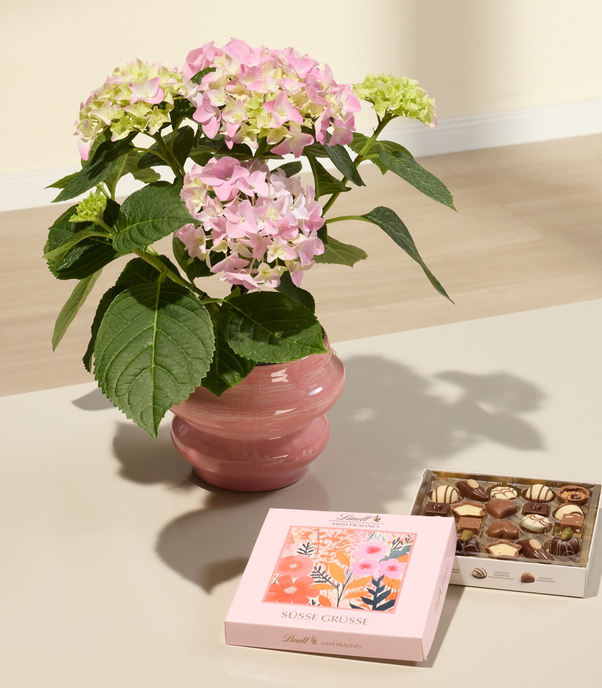 Das Bild zeigt einen Strauß leuchtend rosa und weißer Hortensienblüten in einer rosa Keramikvase, begleitet von einer Schachtel verschiedener Pralinen und einer Grußkarte auf schlichtem weißem Hintergrund.