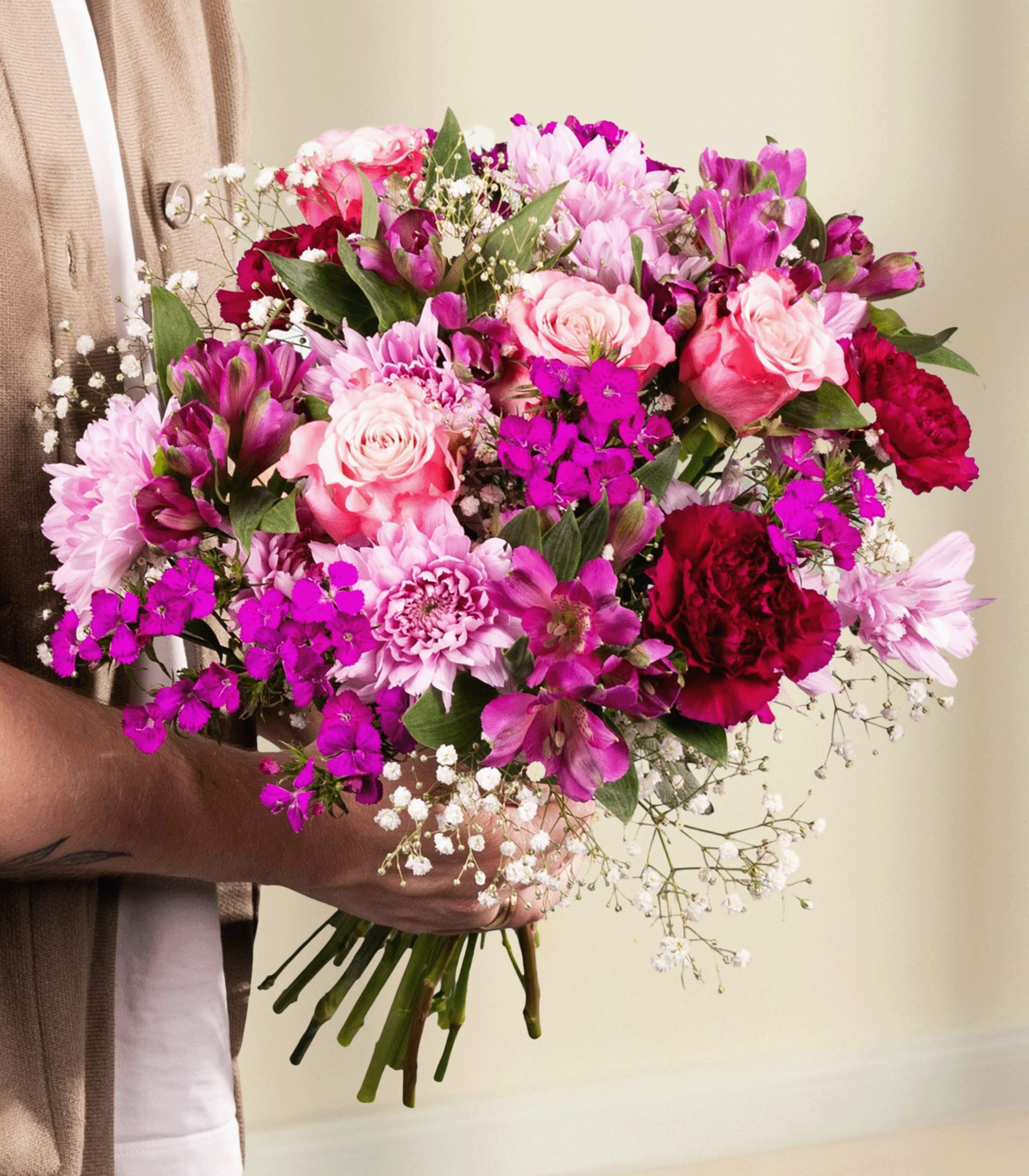Ein leuchtender Strauß rosa, roter und violetter Blumen, gehalten von einer Person in einem beigen Mantel vor einem hellblauen Hintergrund.