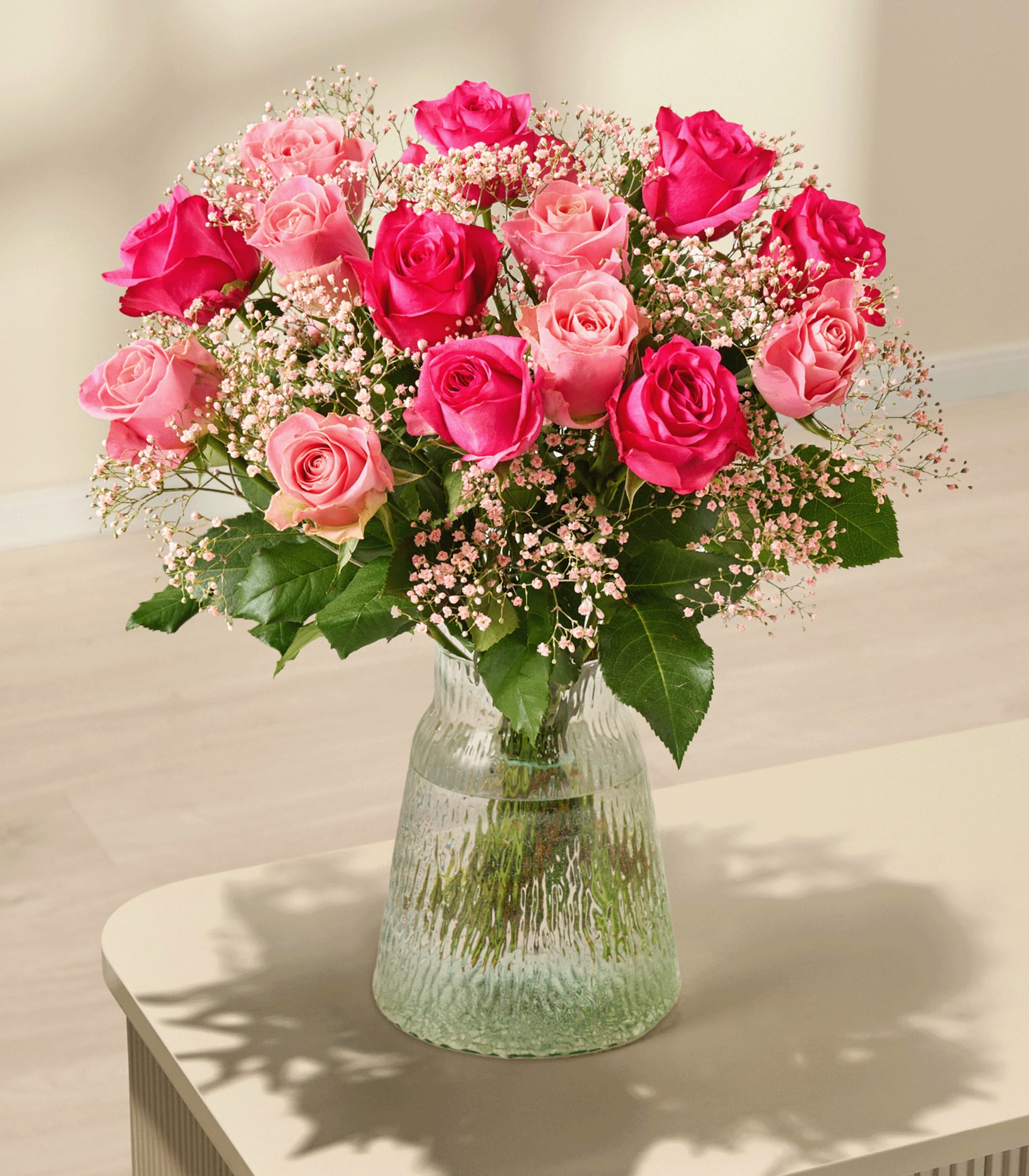 Ein leuchtender Strauß aus rosa und roten Rosen, verziert mit zartem Schleierkraut, wird von einer Person vor einem schlichten weißen Hintergrund in den Händen gehalten.