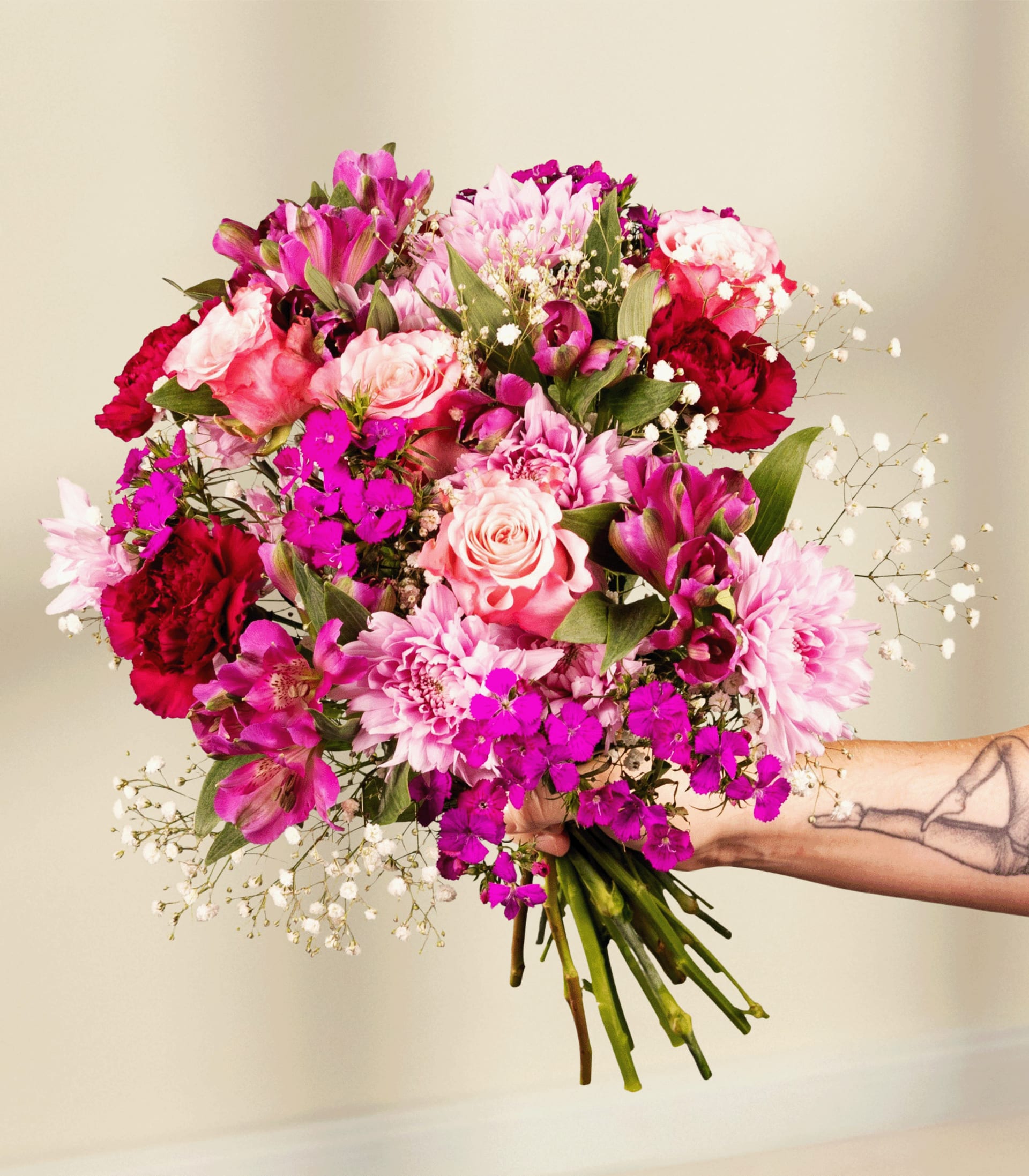 Ein leuchtender Strauß rosa, roter und violetter Blumen, darunter Rosen, Nelken und Schleierkraut, wird von einer tätowierten Hand einer Person vor einem schlichten blauen Hintergrund gehalten.