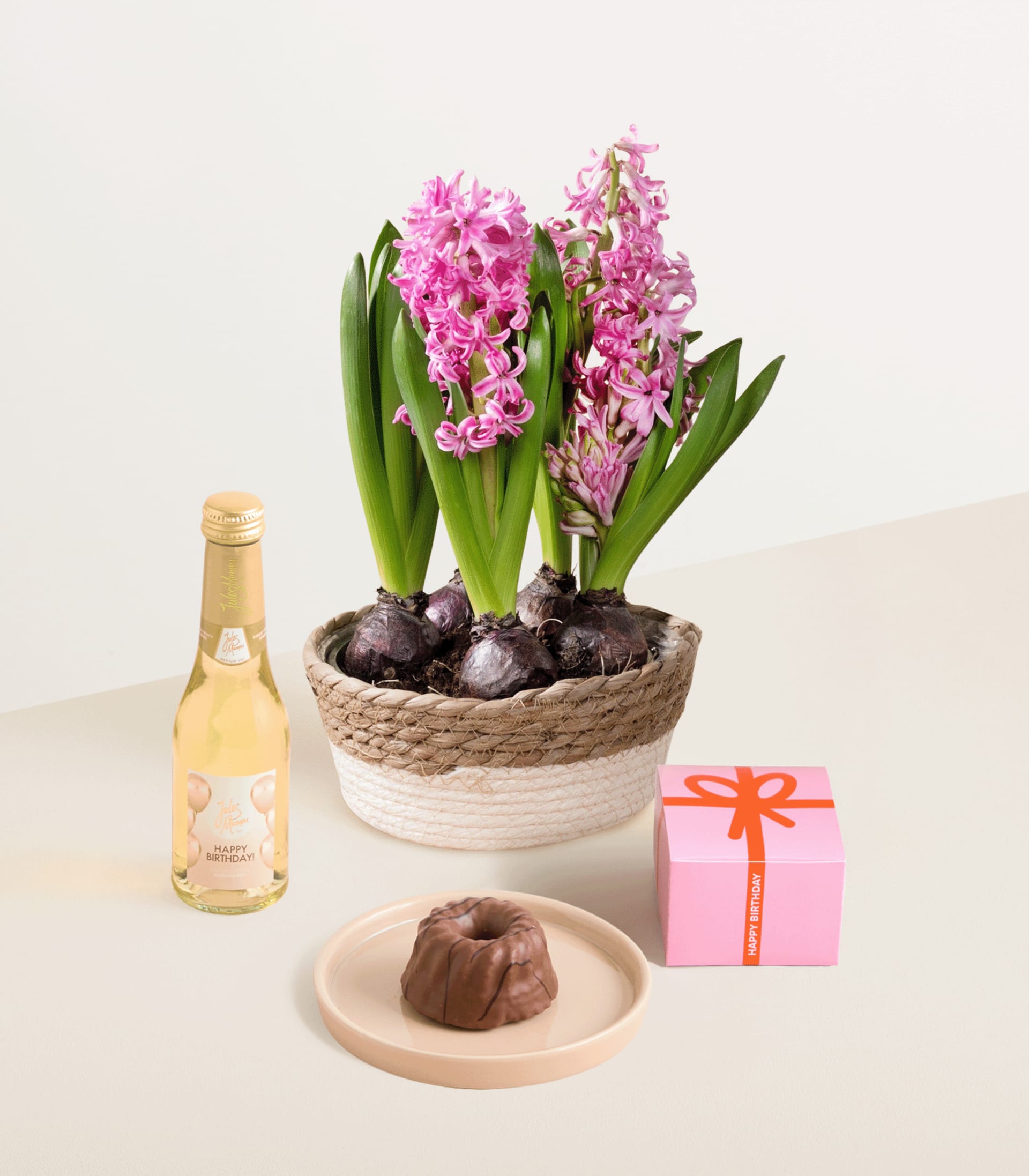 Das Bild zeigt eine Topfpflanze mit leuchtend rosa Hyazinthenblüten, umgeben von einem geflochtenen Korb, einer Flasche, die wie Champagner aussieht, und einem kleinen Teller mit Schokoladenkonfekt.