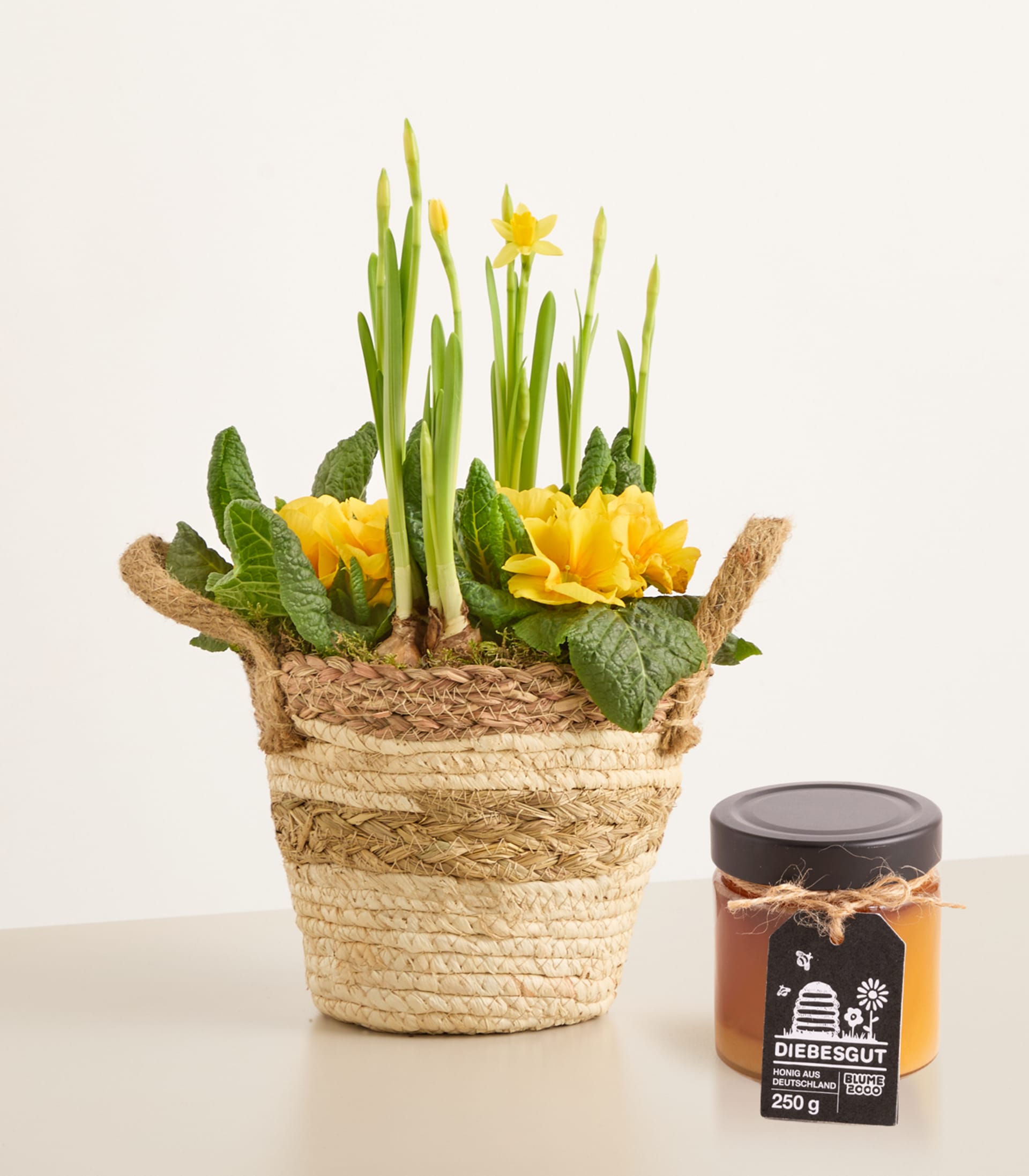 Auf einer Holzoberfläche steht ein geflochtener Korb voller leuchtend rosa und roter Blumen, daneben ein kleines Glas mit etwas, das wie ein Gewürz oder Brotaufstrich aussieht.