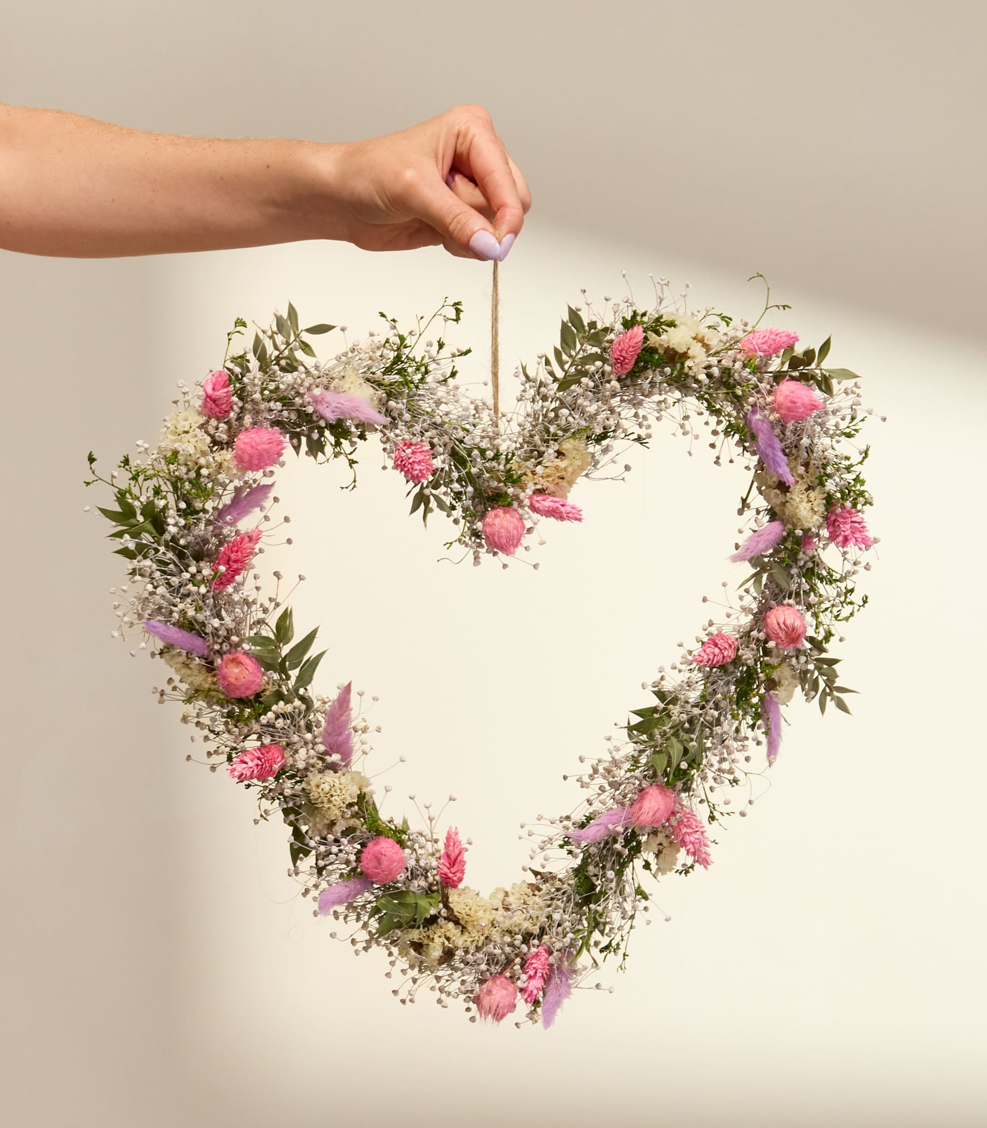 Ein herzförmiger Kranz aus zarten rosa und weißen Blüten wird von einer Hand vor einem schlichten weißen Hintergrund hochgehalten.