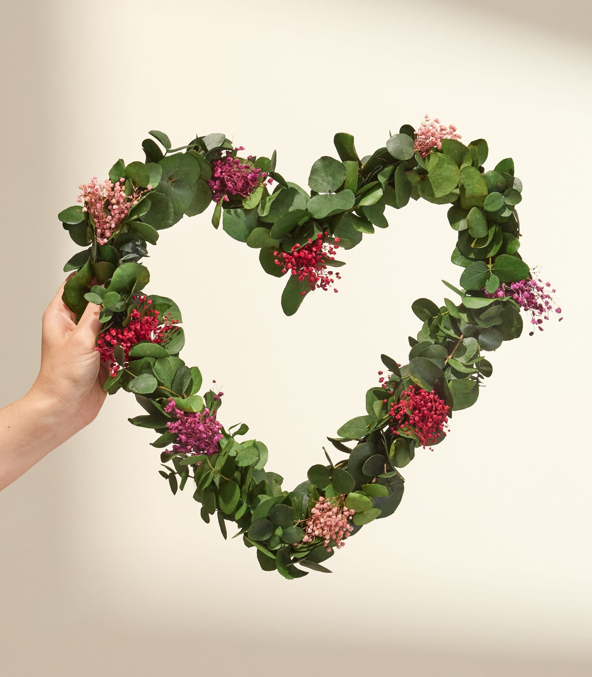 Ein herzförmiger Kranz aus üppigem Grün und leuchtend rosa und roten Blumen wird vor einem schlichten, hellen Hintergrund hochgehalten.