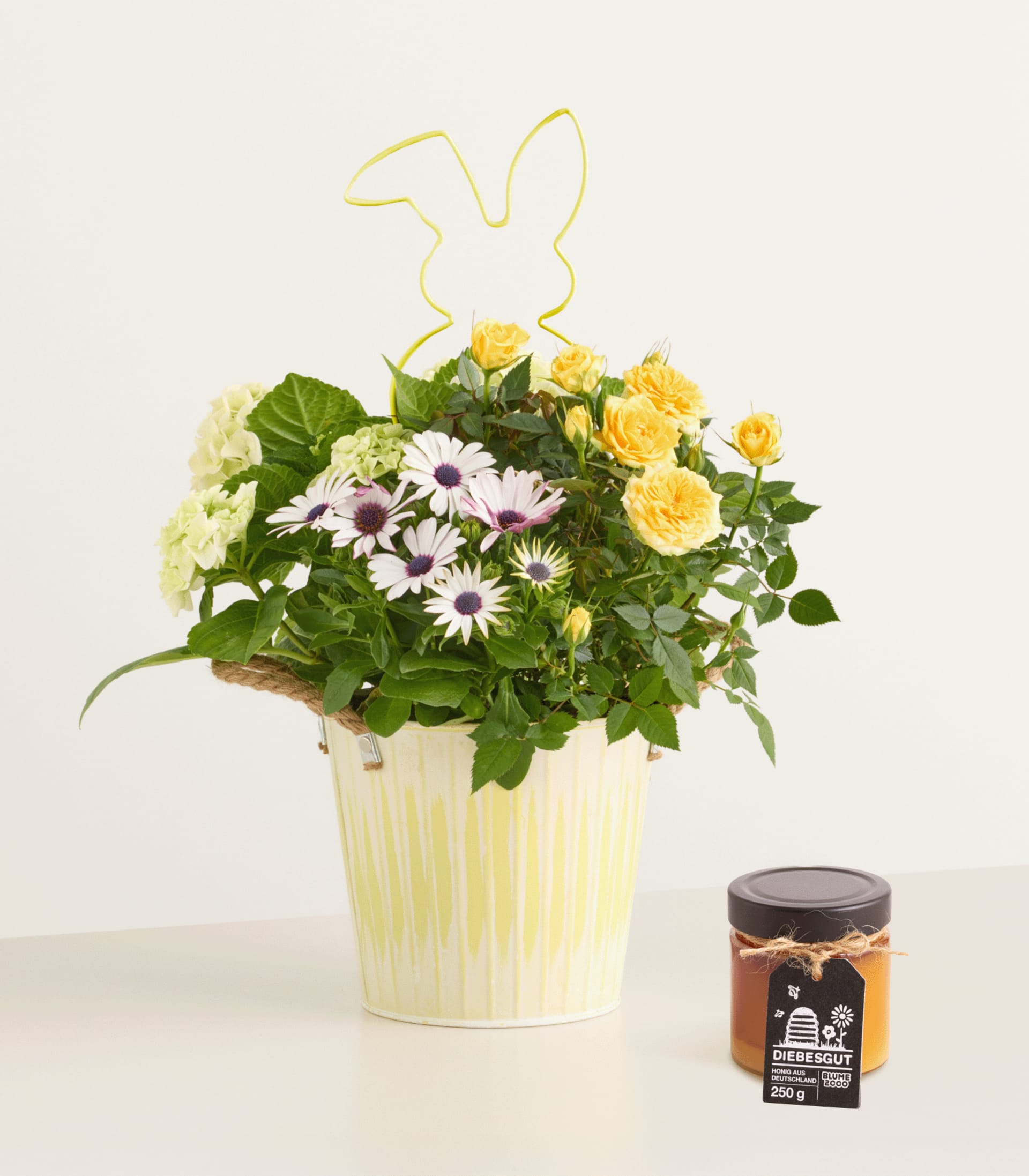 Ein bunter Strauß gelber Rosen, weißer Gänseblümchen und anderer Blumen in einem gelben Keramiktopf, mit einem kleinen Glas mit etwas, das wie Marmelade oder Konfitüre aussieht, im Vordergrund, vor einem schlichten weißen Hintergrund.