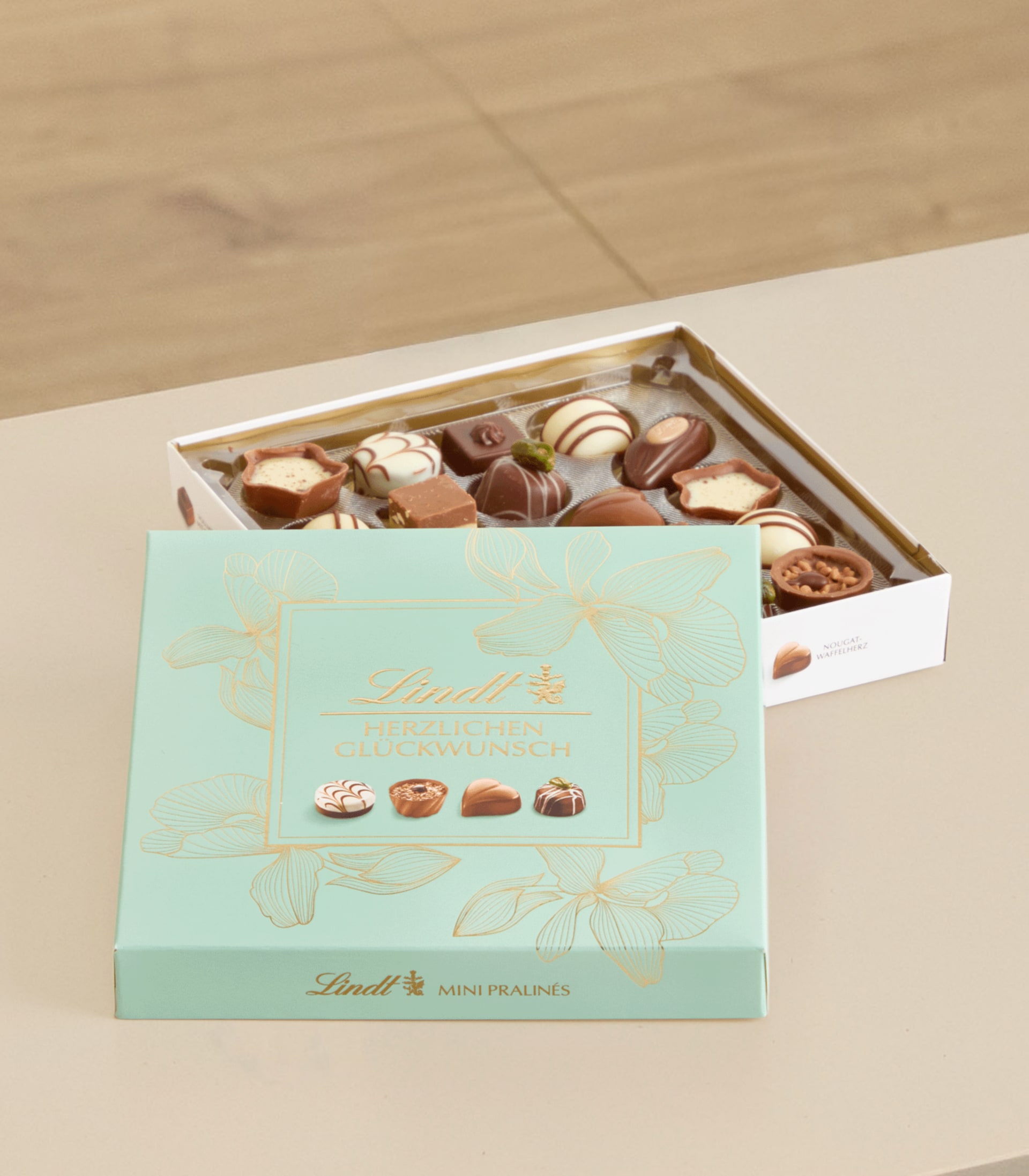 Eine Schachtel mit verschiedenen Lindt-Schokoladen in einer mintgrünen und goldenen Verpackung, die auf einer hellen Oberfläche liegt.