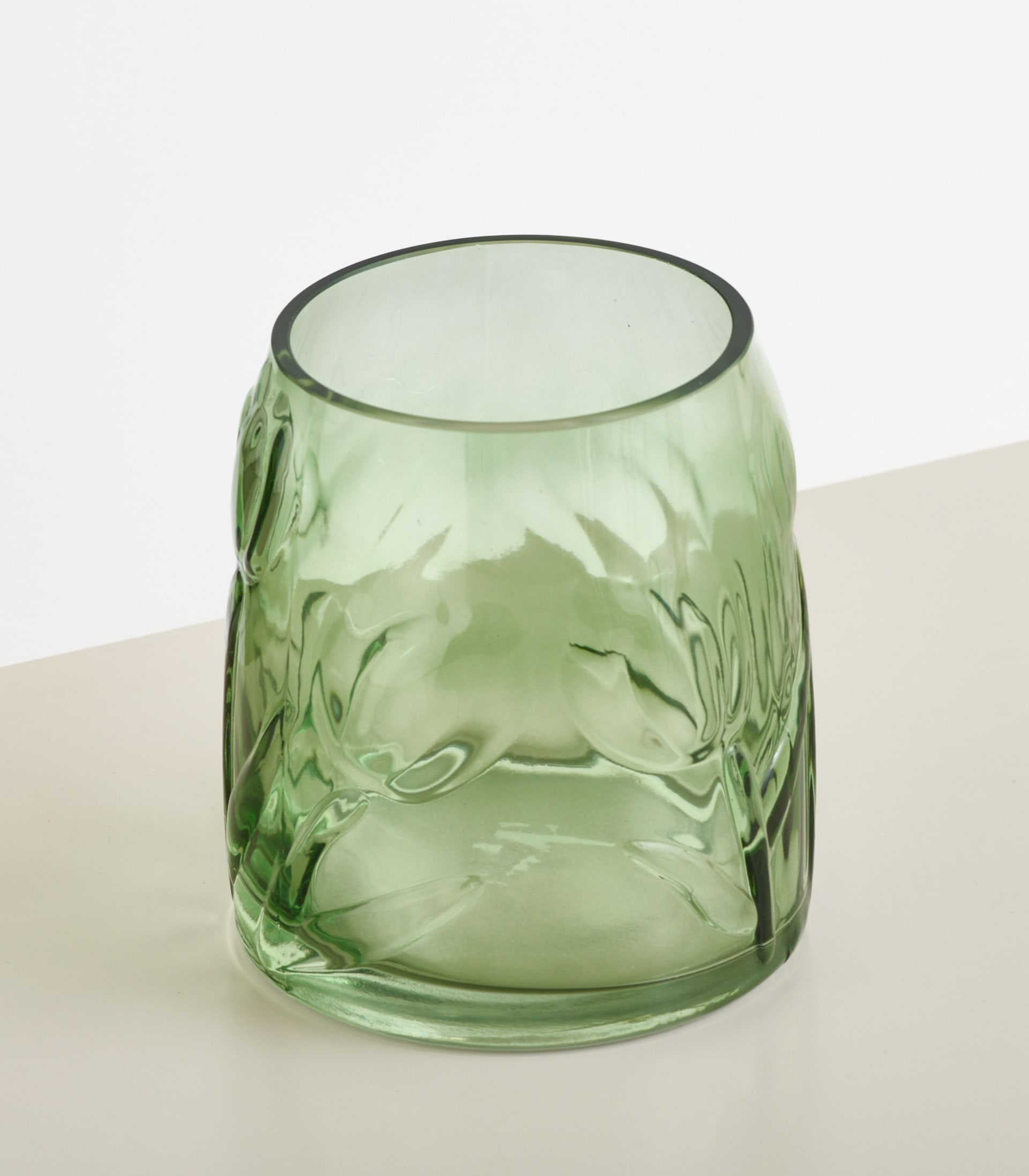 Eine durchscheinende grüne Glasvase mit geprägtem Blumenmuster steht auf einer weißen Fläche vor einem sanft beleuchteten, cremefarbenen Hintergrund.