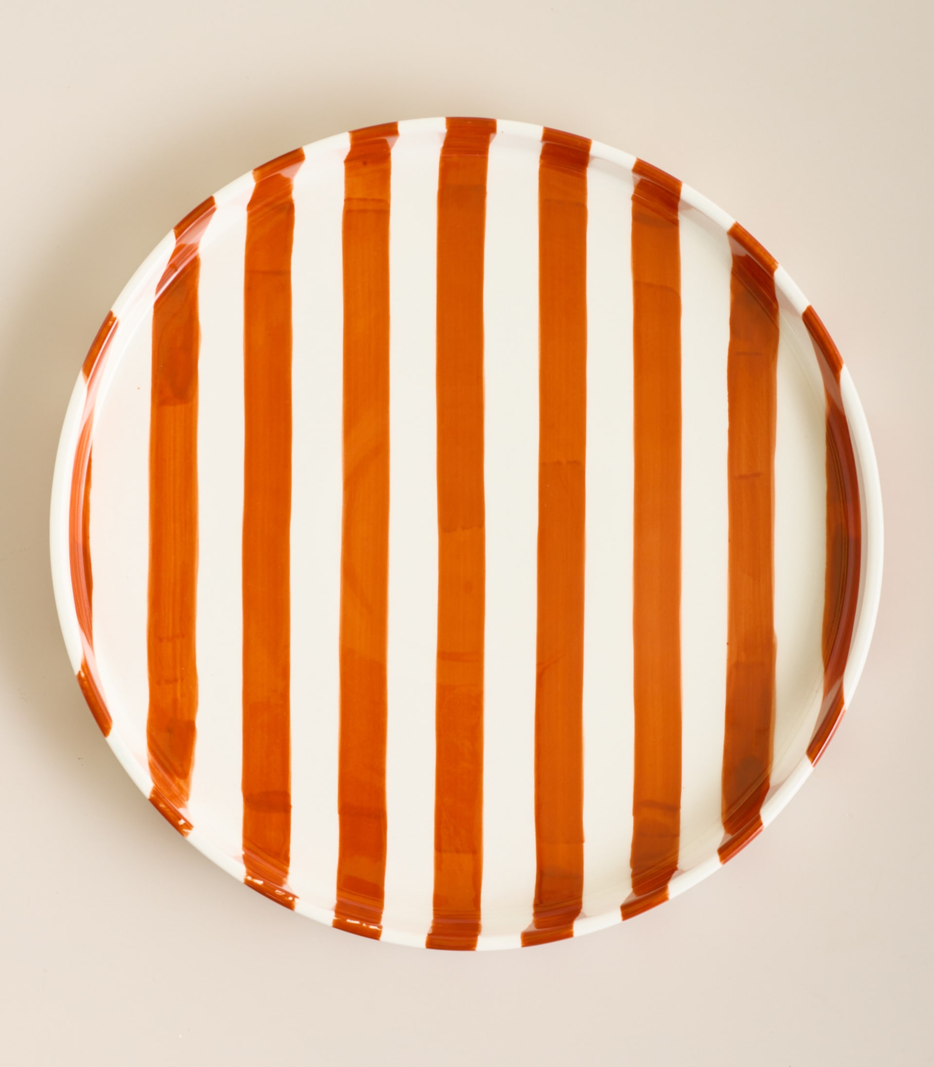 Ein runder Keramikteller mit kräftigen orangefarbenen und weißen vertikalen Streifen steht auf einer schlichten, hellen Oberfläche.
