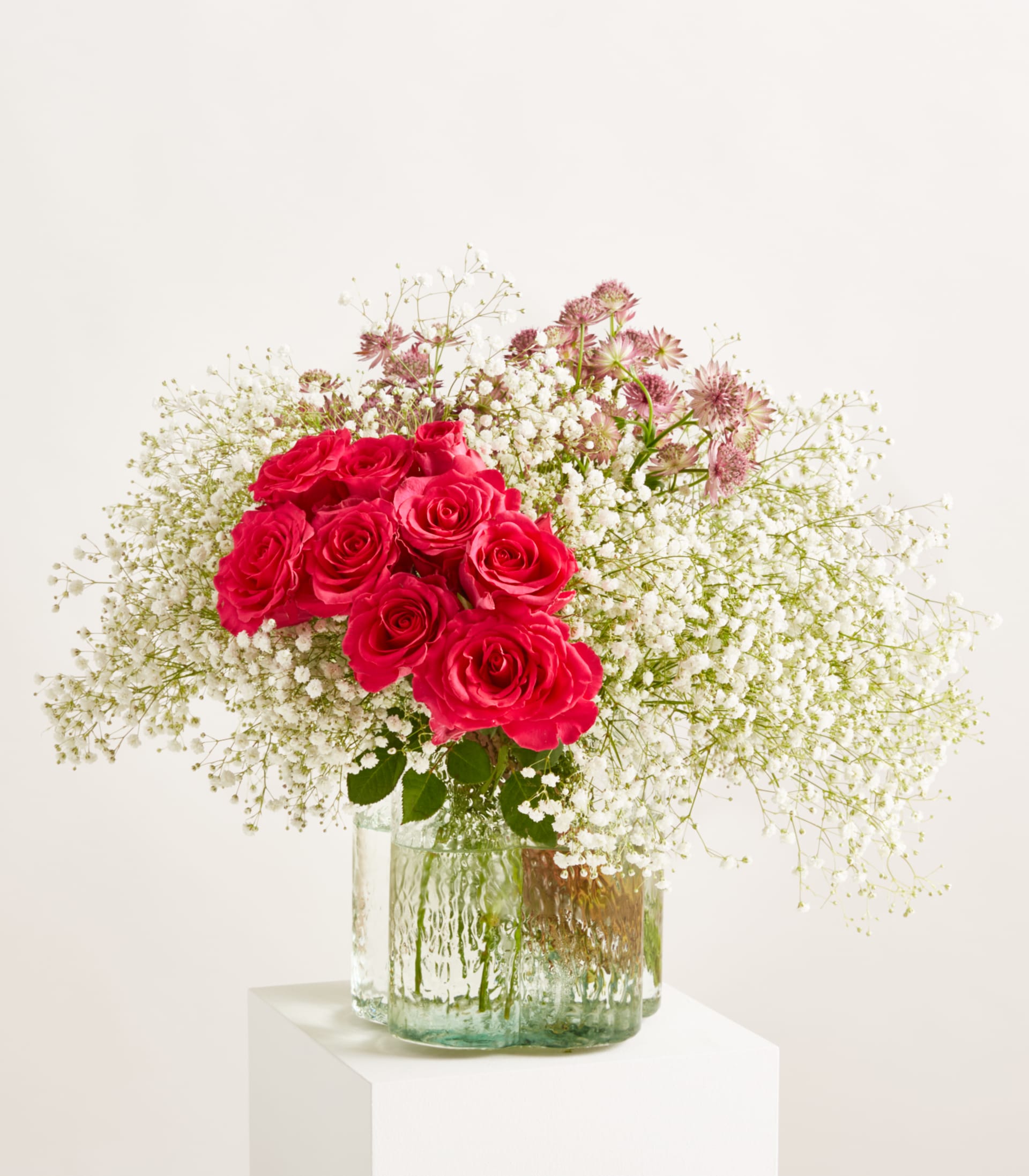 Ein leuchtender Strauß roter Rosen und zartem weißen Schleierkraut ist in einer durchsichtigen Glasvase vor einem sanften, schlichten Hintergrund arrangiert.