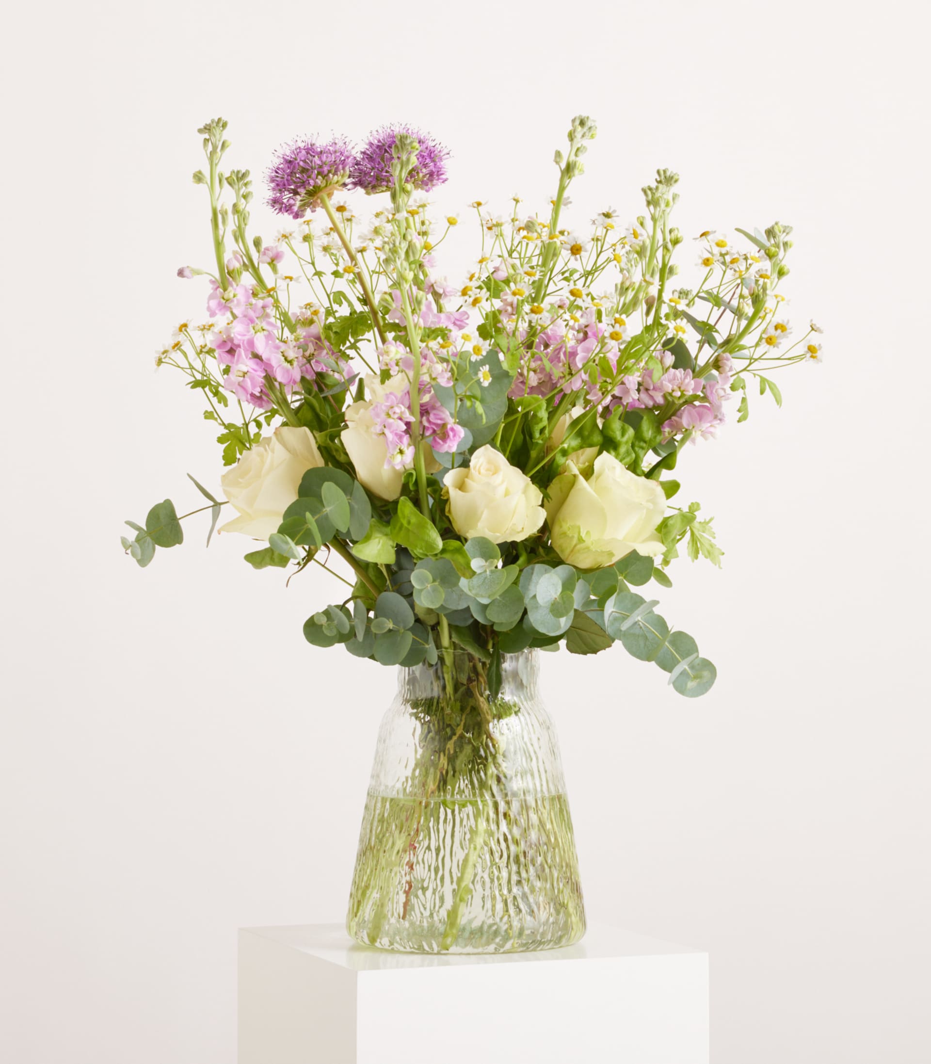 Ein Strauß weißer Rosen und zarter lila und rosa Wildblumen steht in einer strukturierten Glasvase auf einem weißen Sockel vor einem schlichten weißen Hintergrund.