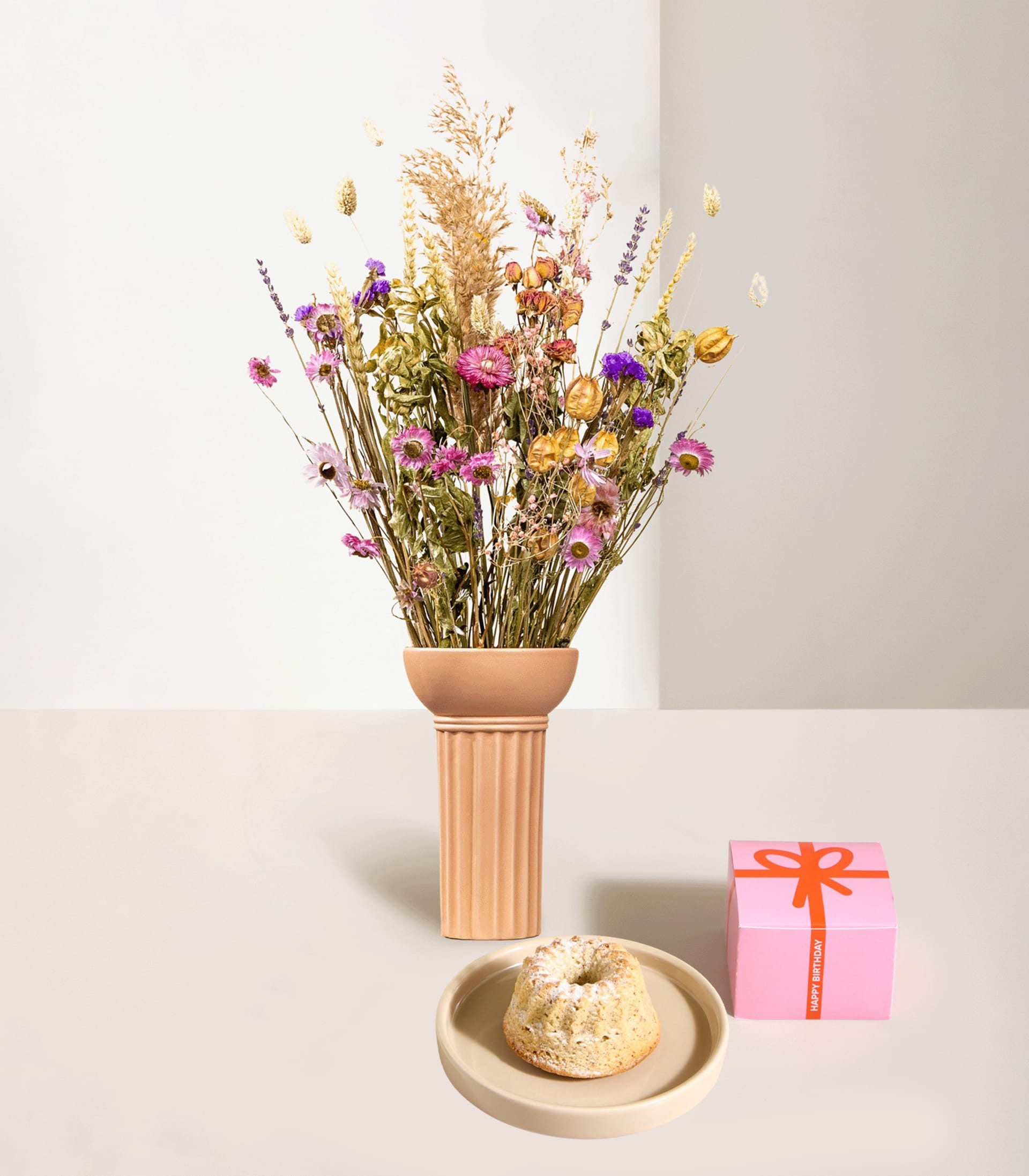 Hinter einem kleinen Kuchen auf einem Teller und einer rosa Geschenkbox steht eine Vase mit Trockenblumen, alles vor einem schlichten Hintergrund.