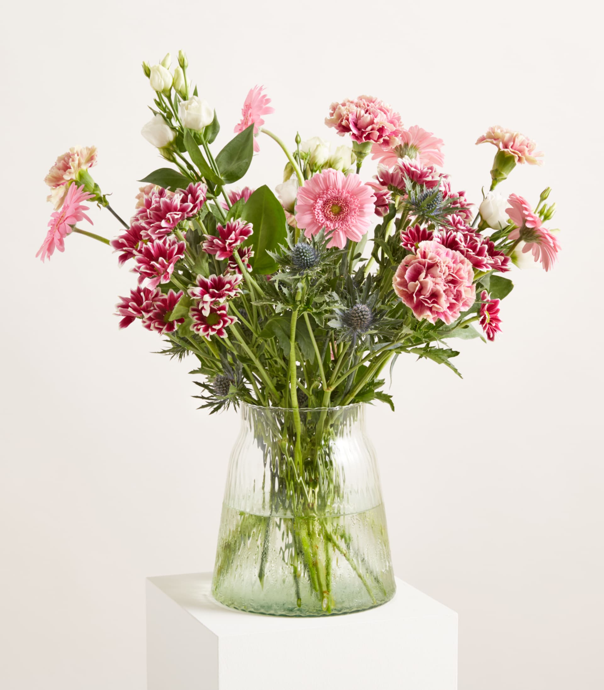 Ein farbenfroher Strauß aus rosa und weißen Blumen, darunter Gerbera und Nelken, ist in einer durchsichtigen, gerippten Glasvase vor einem schlichten weißen Hintergrund arrangiert.