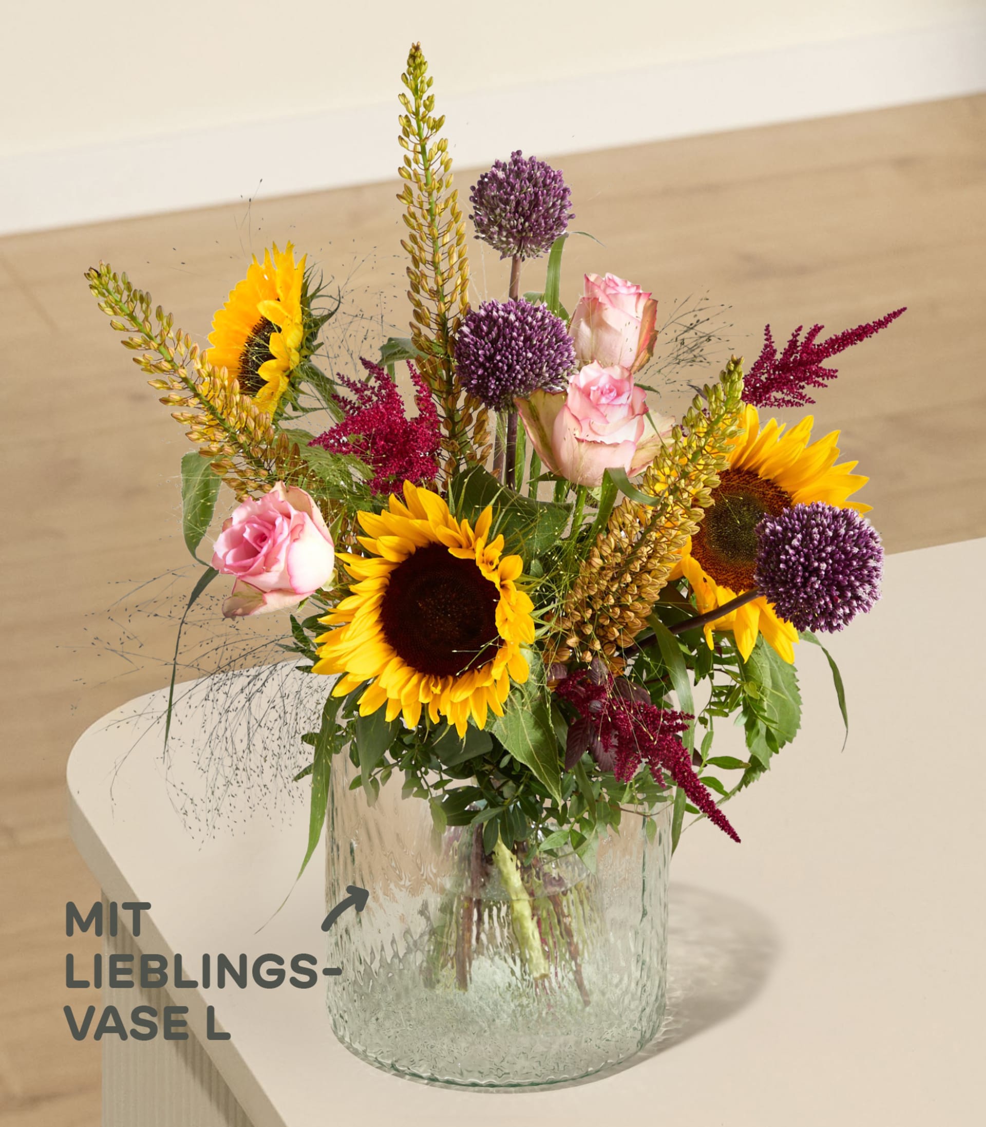 Ein farbenfroher Strauß aus Sonnenblumen, Rosen und lila Zierlauch steht in einer strukturierten Glasvase auf einer hellen Oberfläche, im Hintergrund sind Wand und Boden verschwommen zu sehen.