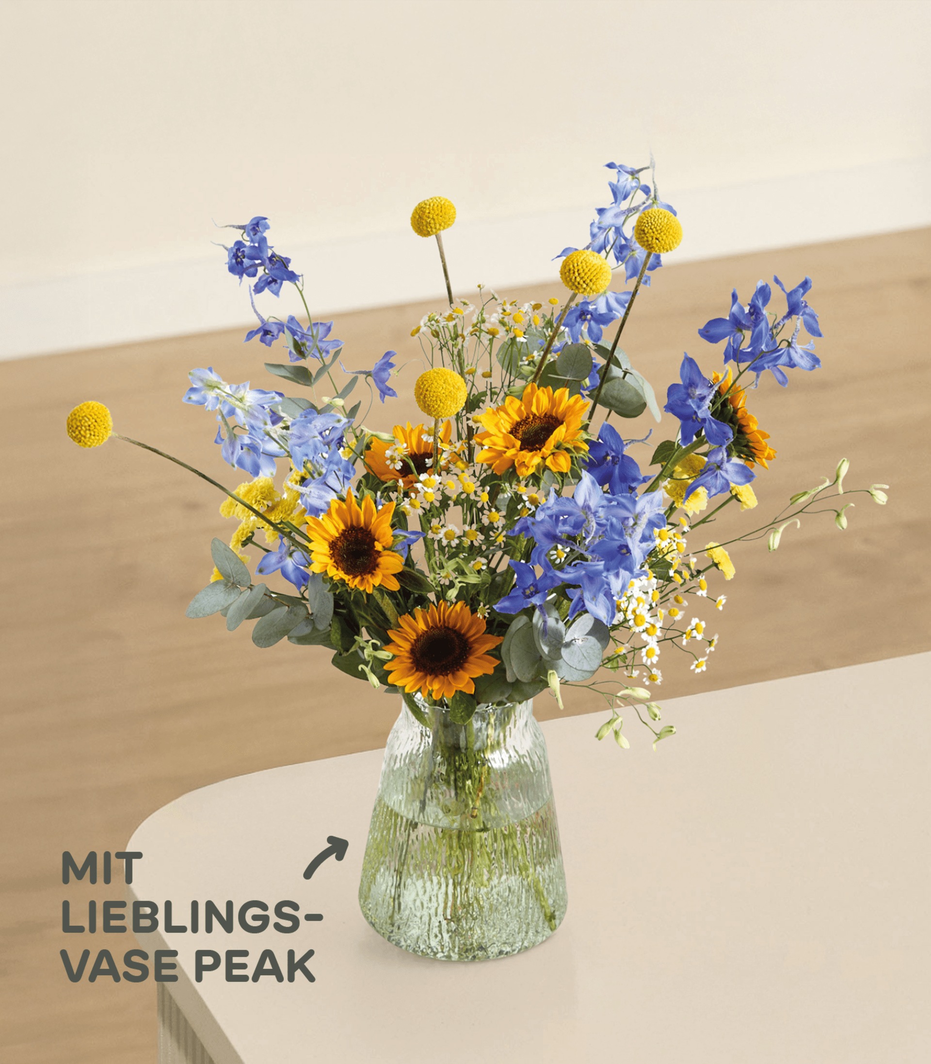Im Vordergrund steht eine strukturierte Glasvase mit einem farbenfrohen Strauß aus Sonnenblumen, blauen Ritterspornen und gelben Knopfblumen vor einem sanft beleuchteten, neutralen Hintergrund mit deutschem Text.