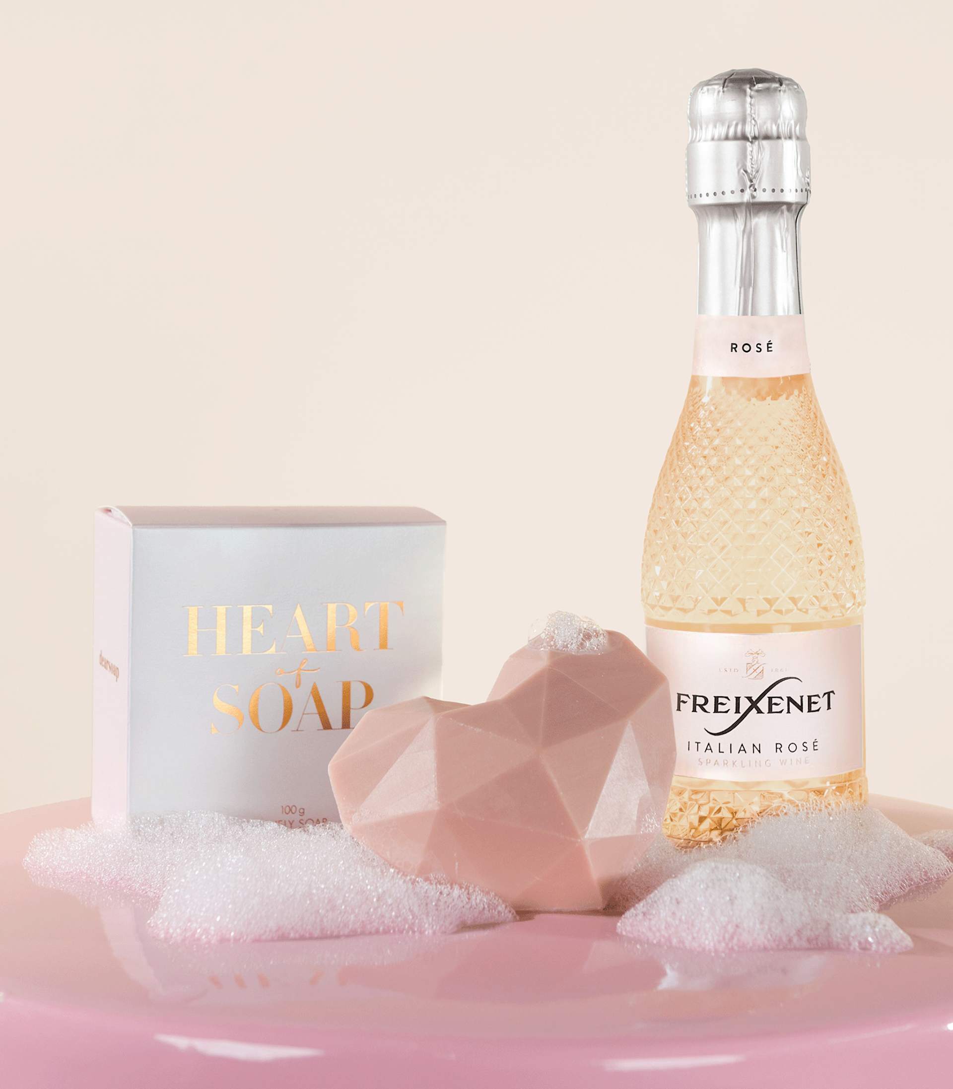 Auf einer weichen, flauschigen rosa Oberfläche stehen eine rosafarbene Glasflasche mit Sekt, eine weiße Schachtel mit der Aufschrift „Luxusseife“ und ein rosafarbenes, geometrisch geformtes Objekt.