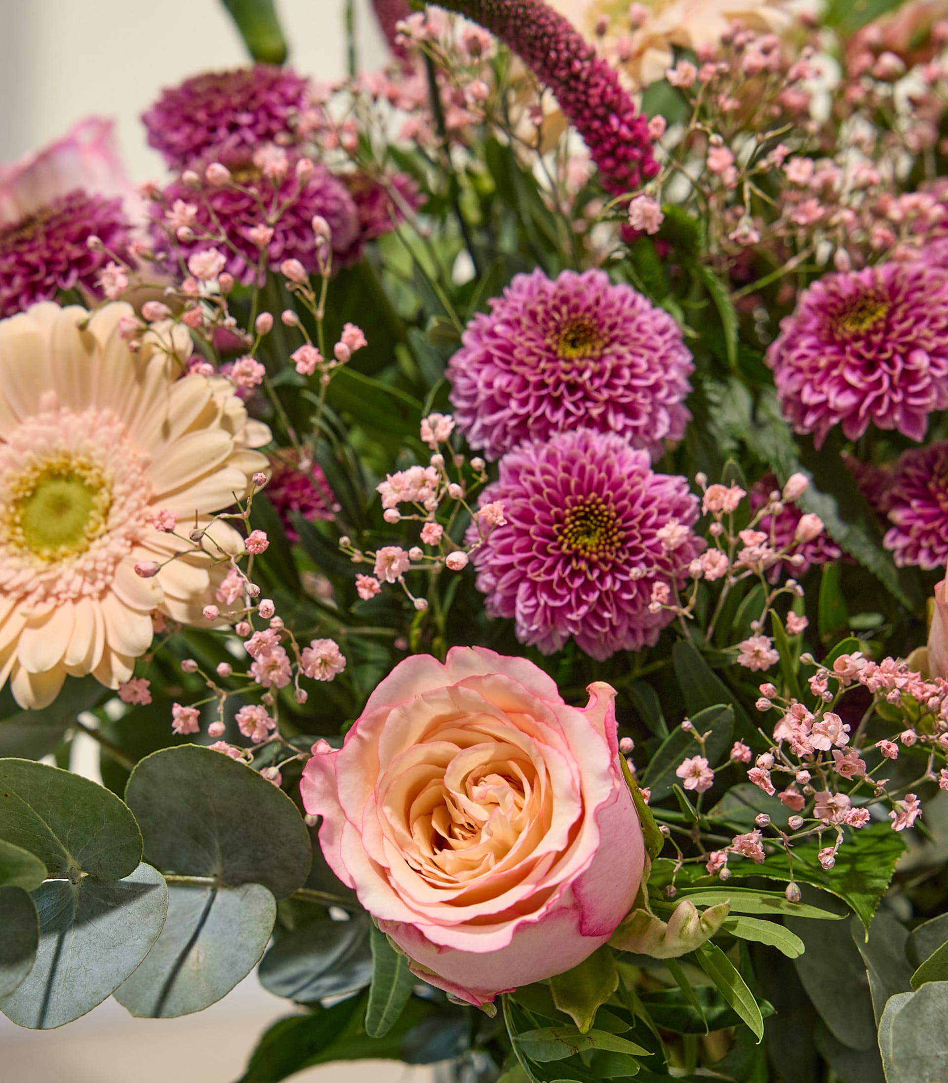 Ein lebhafter Strauß aus verschiedenen rosa und gelben Blumen, darunter Rosen, Gerbera und andere zarte Blüten, arrangiert vor einem schlichten weißen Hintergrund.