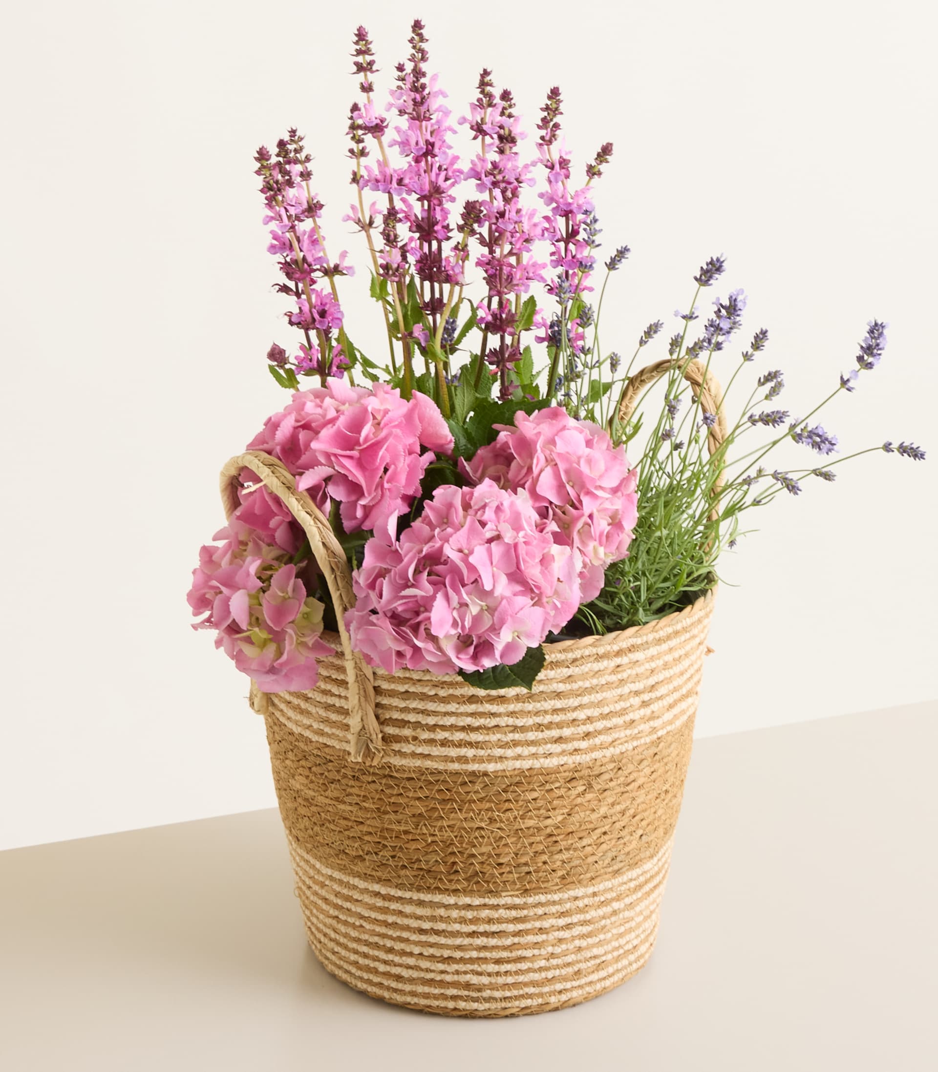 Ein geflochtener Korb, gefüllt mit rosa Hortensien und lila Blüten, steht auf einer neutralen Fläche vor einem schlichten Hintergrund.