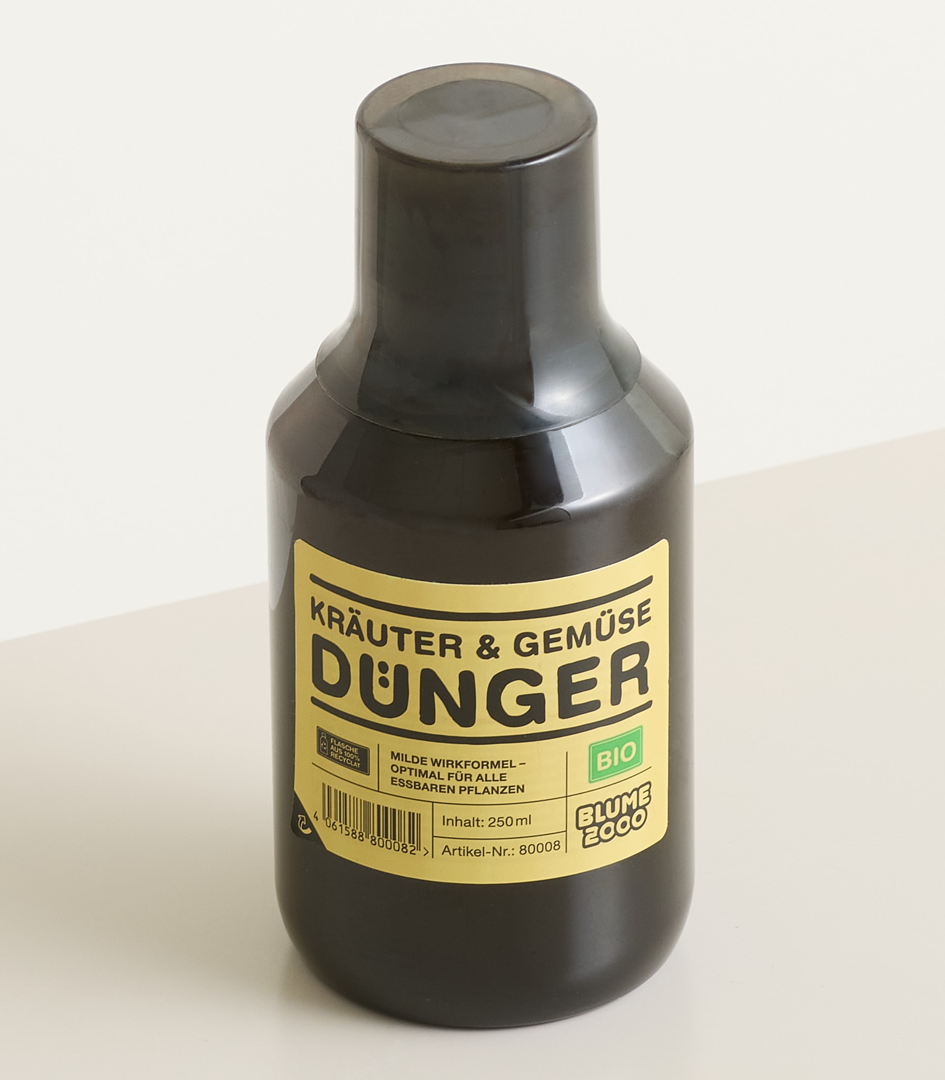 Im Vordergrund steht vor einem schlichten weißen Hintergrund eine dunkelbraune Flasche „Kräuter & Gemüse Dünger“ mit gelbem Etikett.