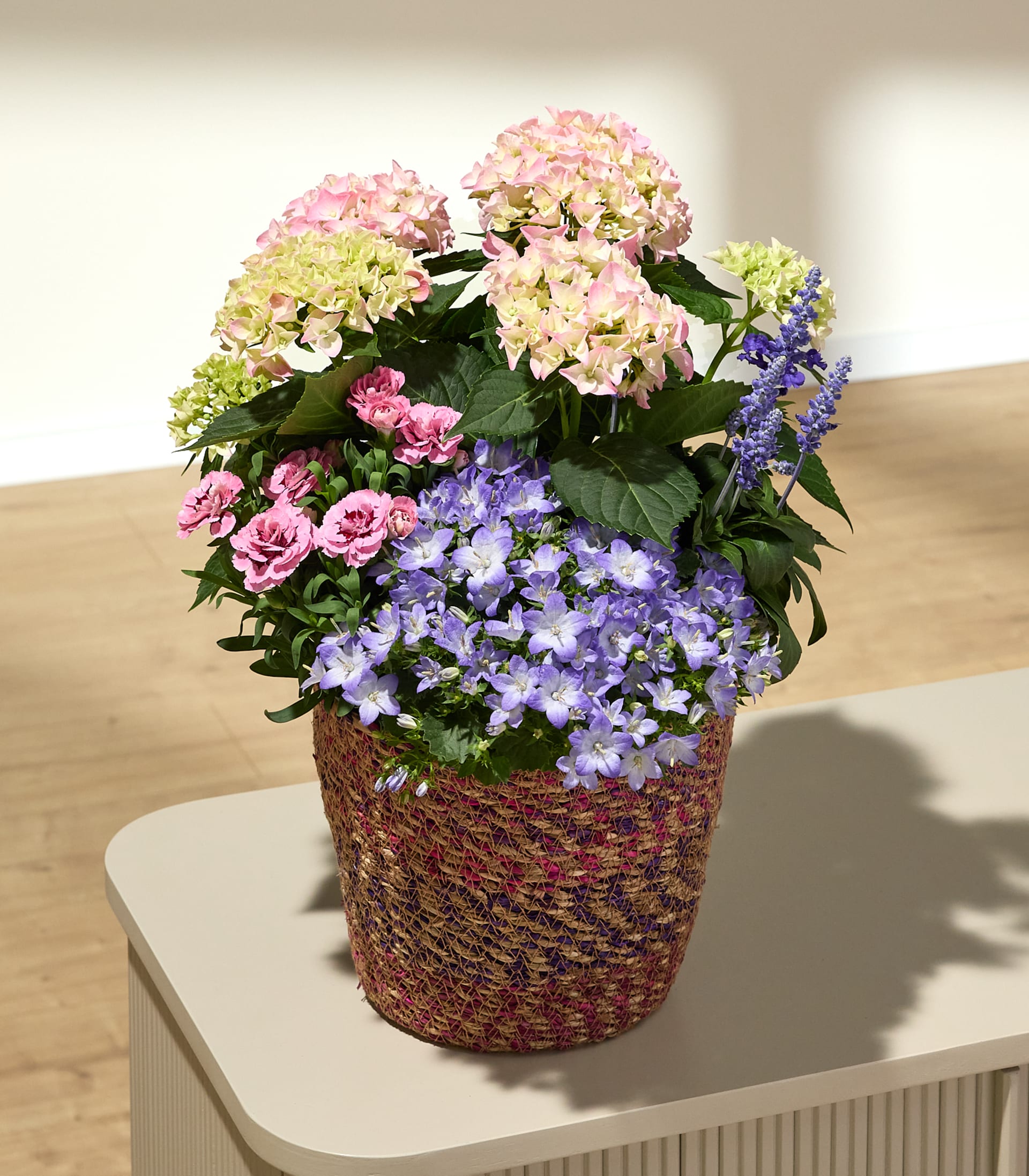 Ein geflochtener Korb, prall gefüllt mit rosa und violetten Blumen, steht vor einer hellen Wand und einem Holzboden.