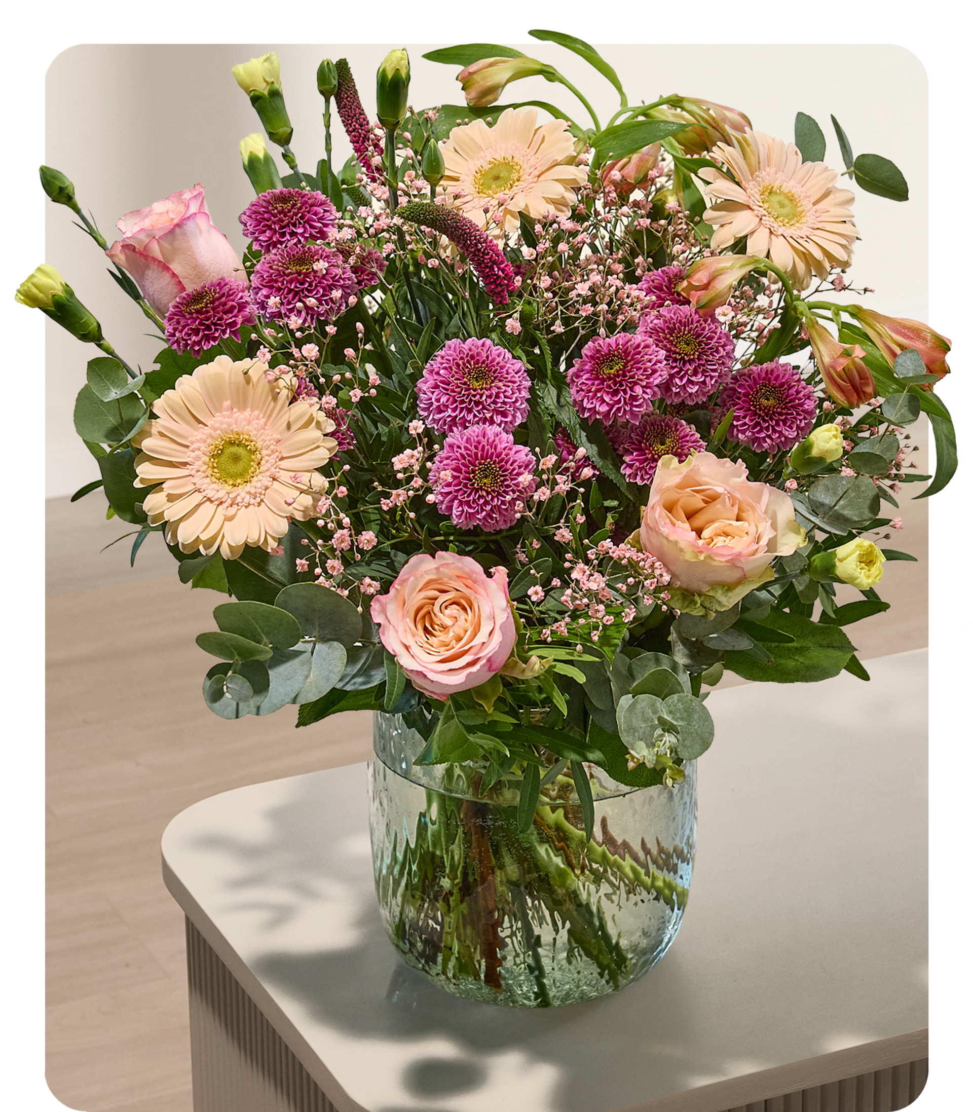 Ein leuchtender Strauß rosa und pfirsichfarbener Blumen füllt eine durchsichtige Glasvase auf einem hellen Tisch vor einem weichen, verschwommenen Hintergrund.
