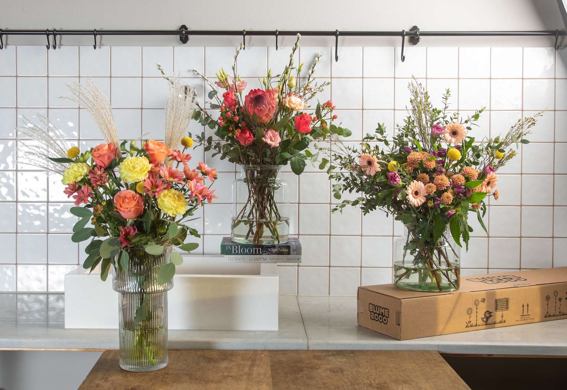 Lebendige Blumenarrangements in Glasvasen schmücken eine geflieste Theke, im Hintergrund steht ein Karton, der eine optisch ansprechende und natürliche Atmosphäre schafft.