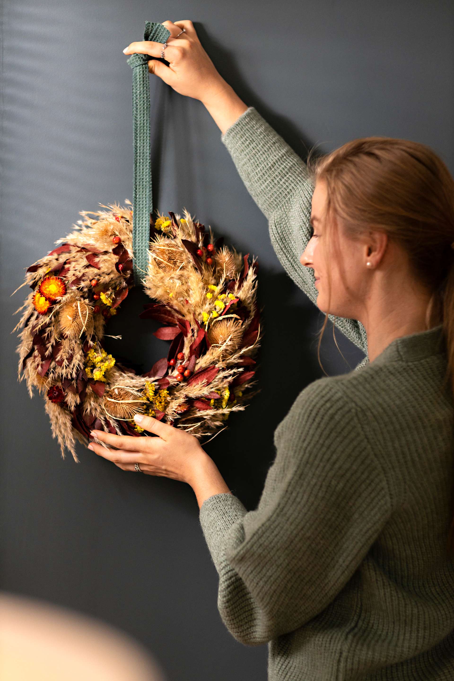 Eine Frau im grünen Pullover hält vor dunklem Hintergrund einen bunten Herbstkranz aus getrockneten Blättern und Blüten.