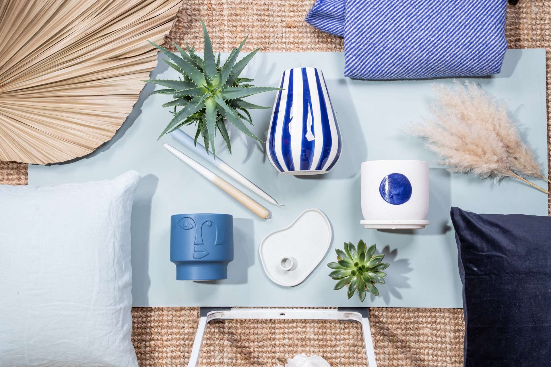 Das Bild zeigt eine Auswahl dekorativer und funktionaler Gegenstände, darunter eine Sukkulente im Topf, eine gestreifte Vase, ein blau-weißes Keramikstück sowie verschiedene Textilien und Accessoires, die auf einer gewebten Matte vor einem neutralen Hintergrund angeordnet sind. (Beschreibungstext KI-generiert)