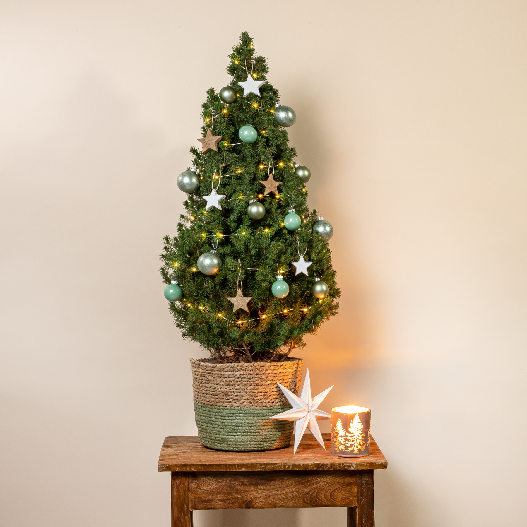 Auf einem Holztisch steht vor einem schlichten beigen Hintergrund ein kleiner, geschmückter Weihnachtsbaum, umgeben von festlichem Schmuck und einem sternförmigen Licht.