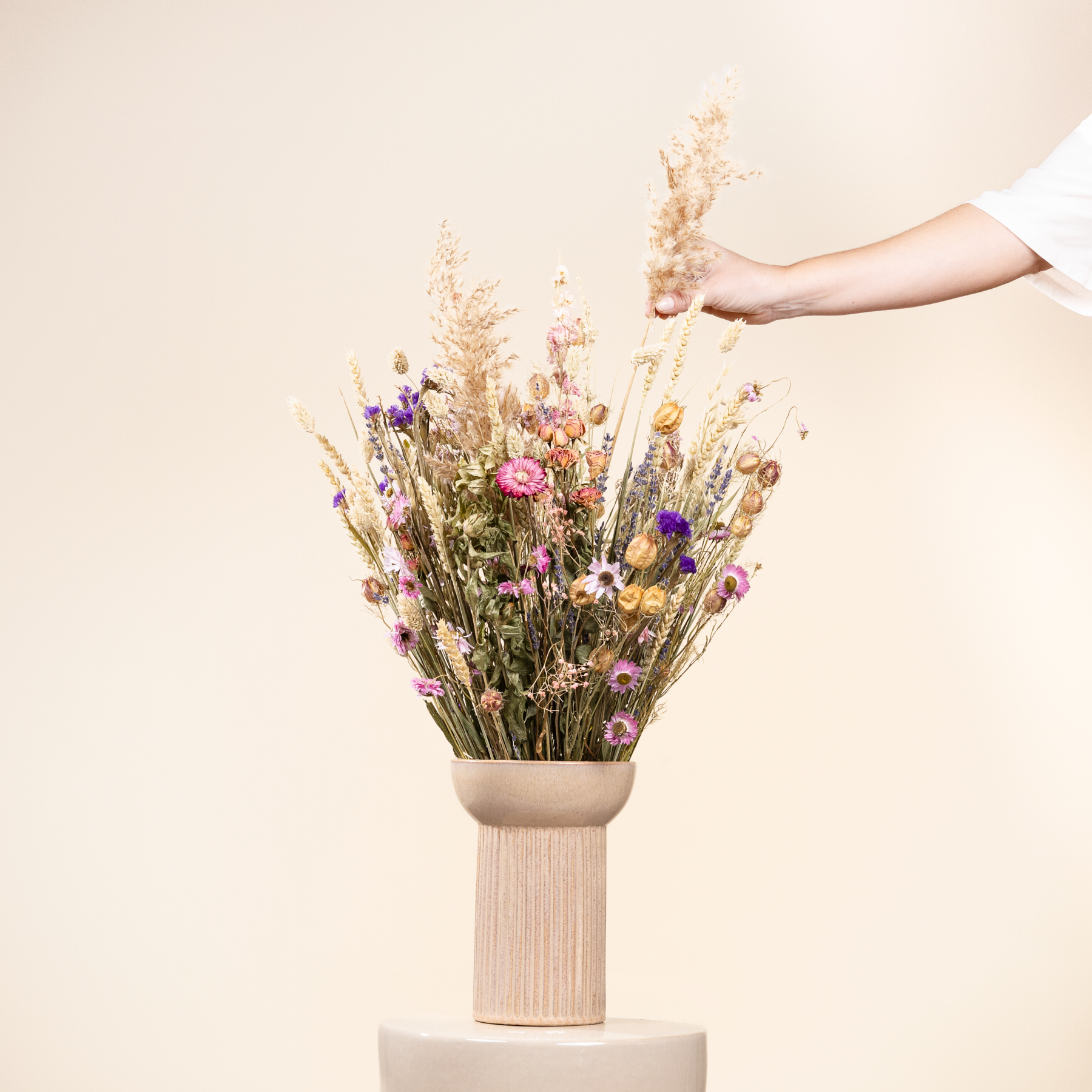 Eine Hand hält eine Vase mit einem farbenfrohen Strauß getrockneter Wildblumen vor einem schlichten, hellen Hintergrund.