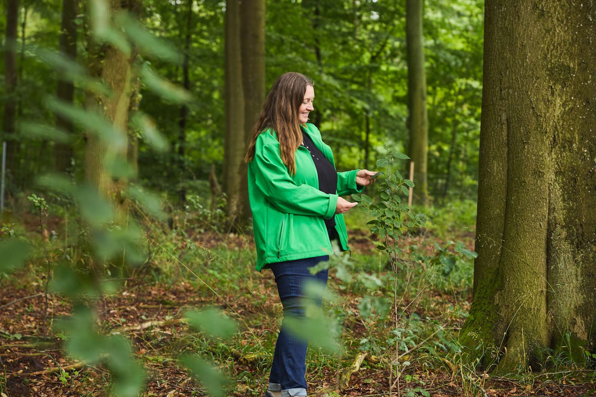 Eine junge Frau in einer grünen Jacke steht in einer üppigen Waldumgebung, umgeben von Bäumen und Grün.