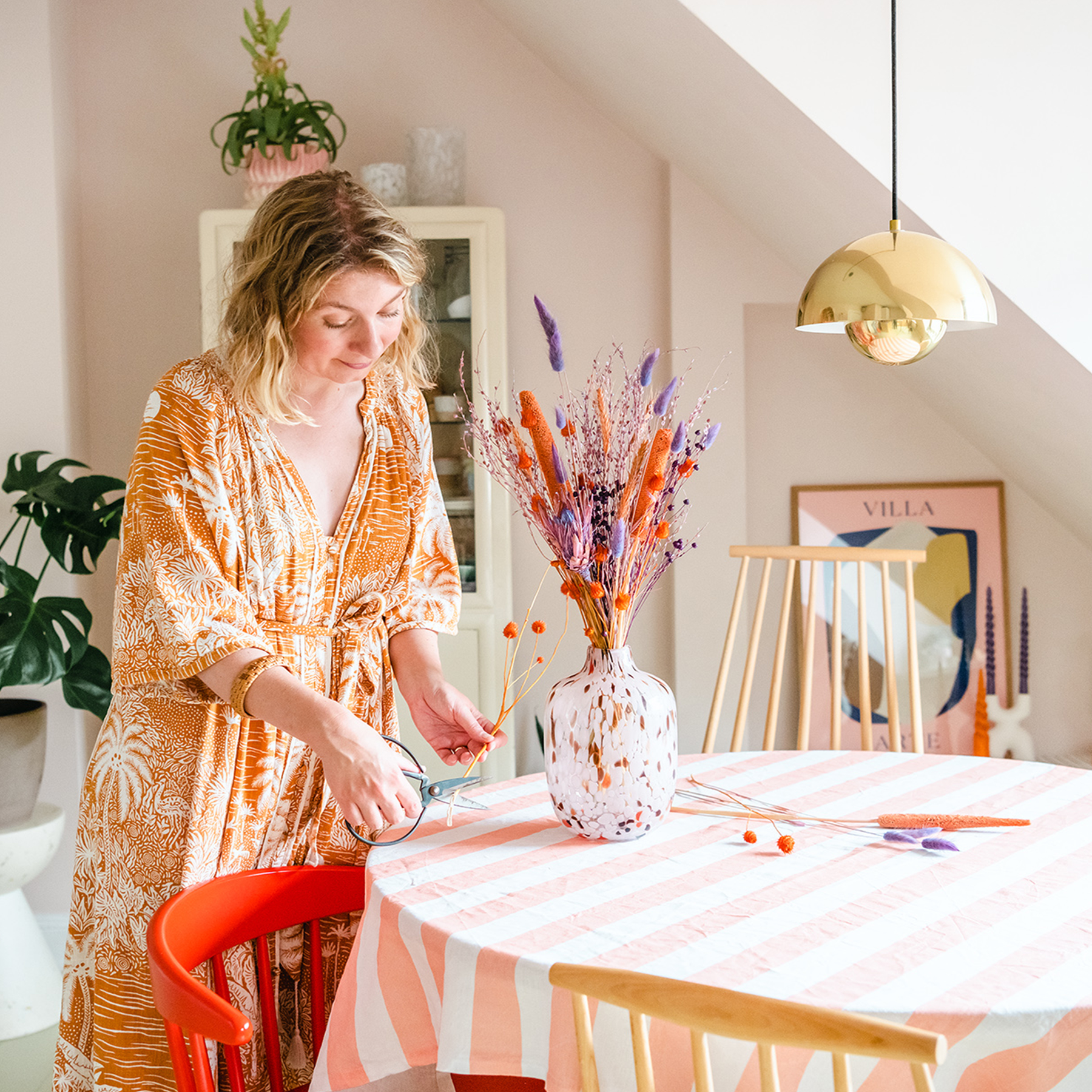 Eine Frau in einem farbenfrohen, gemusterten Kleid sitzt an einem Tisch, der mit einer Vase mit Trockenblumen geschmückt ist, umgeben von einem gemütlichen, mit Pflanzen gefüllten Interieur mit warmer Beleuchtung.