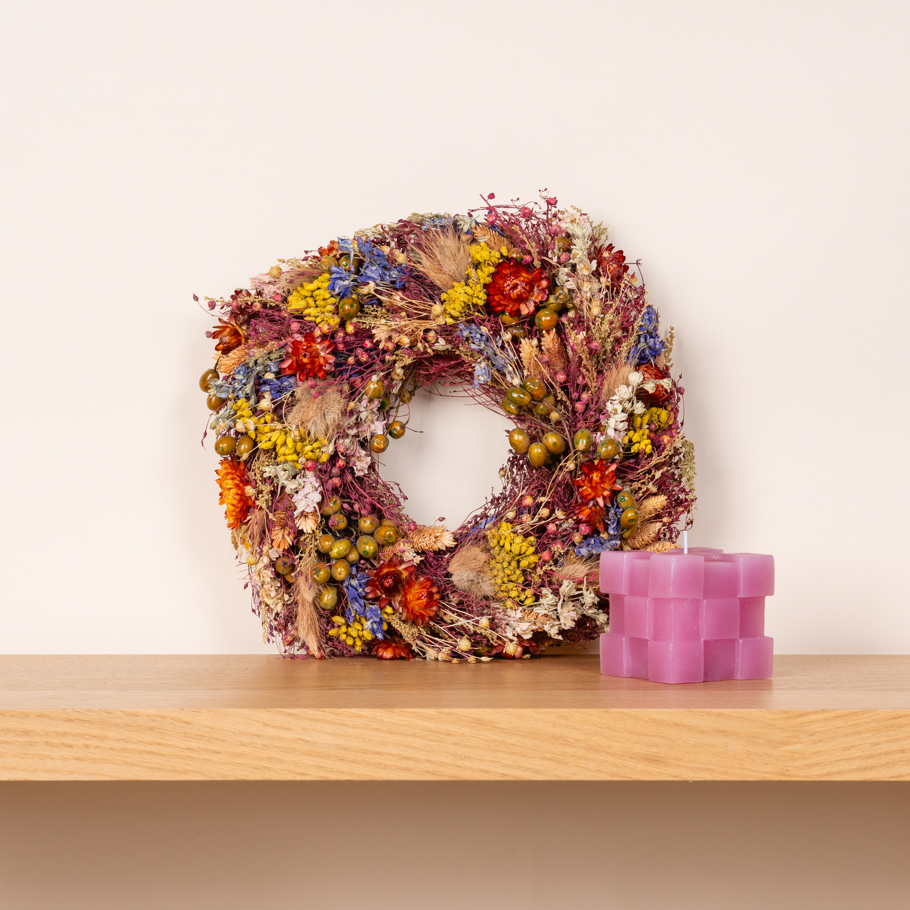 Ein farbenfroher Kranz aus getrockneten Blumen schmückt ein Holzregal, begleitet von einer rosa geometrischen Skulptur.
