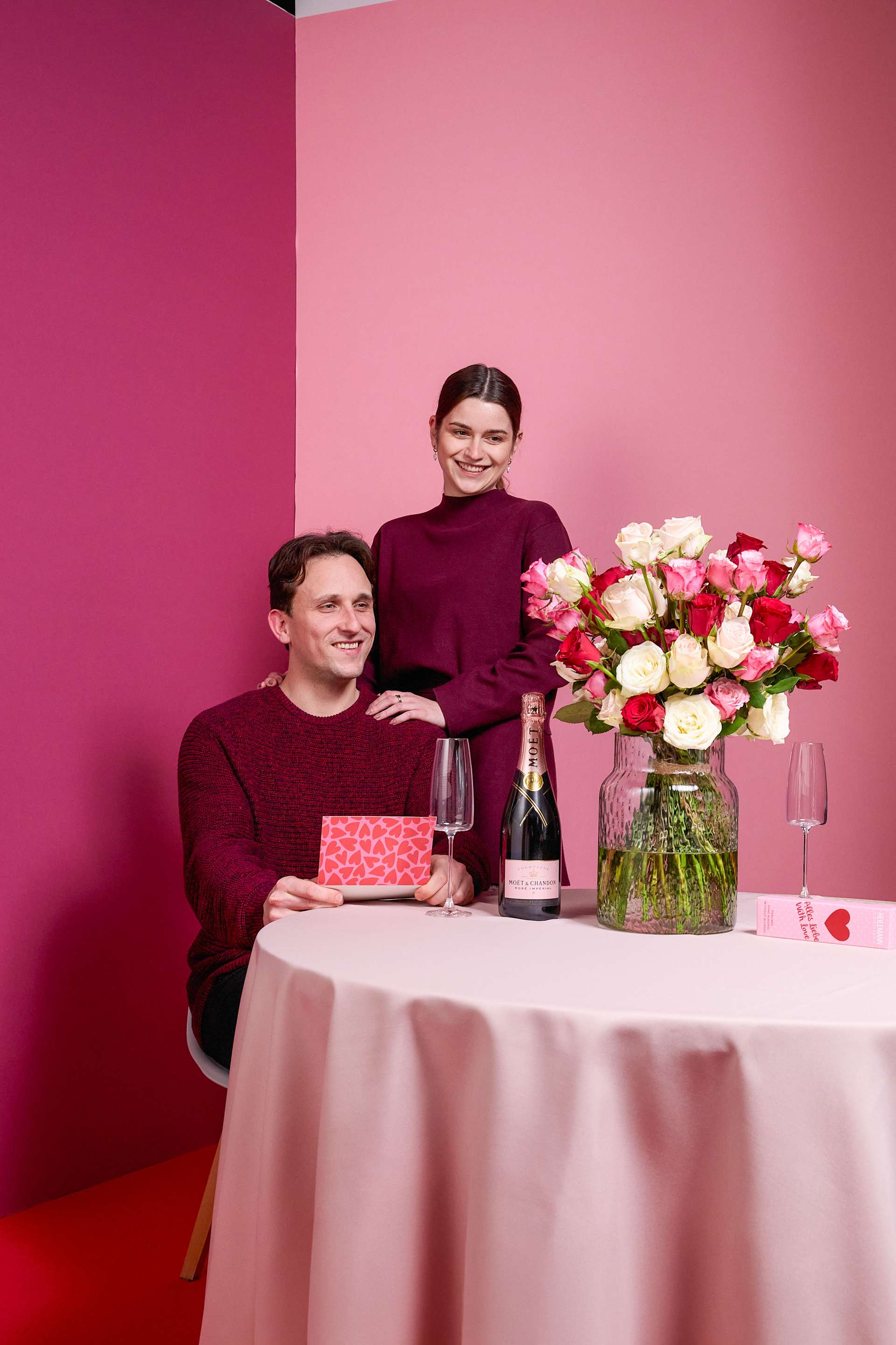 Ein Paar in passenden burgunderfarbenen Pullovern sitzt mit einem Strauß Rosen, einer Flasche Champagner und einer Geschenkbox vor einer leuchtend rosa Wand im Hintergrund an einem Tisch.