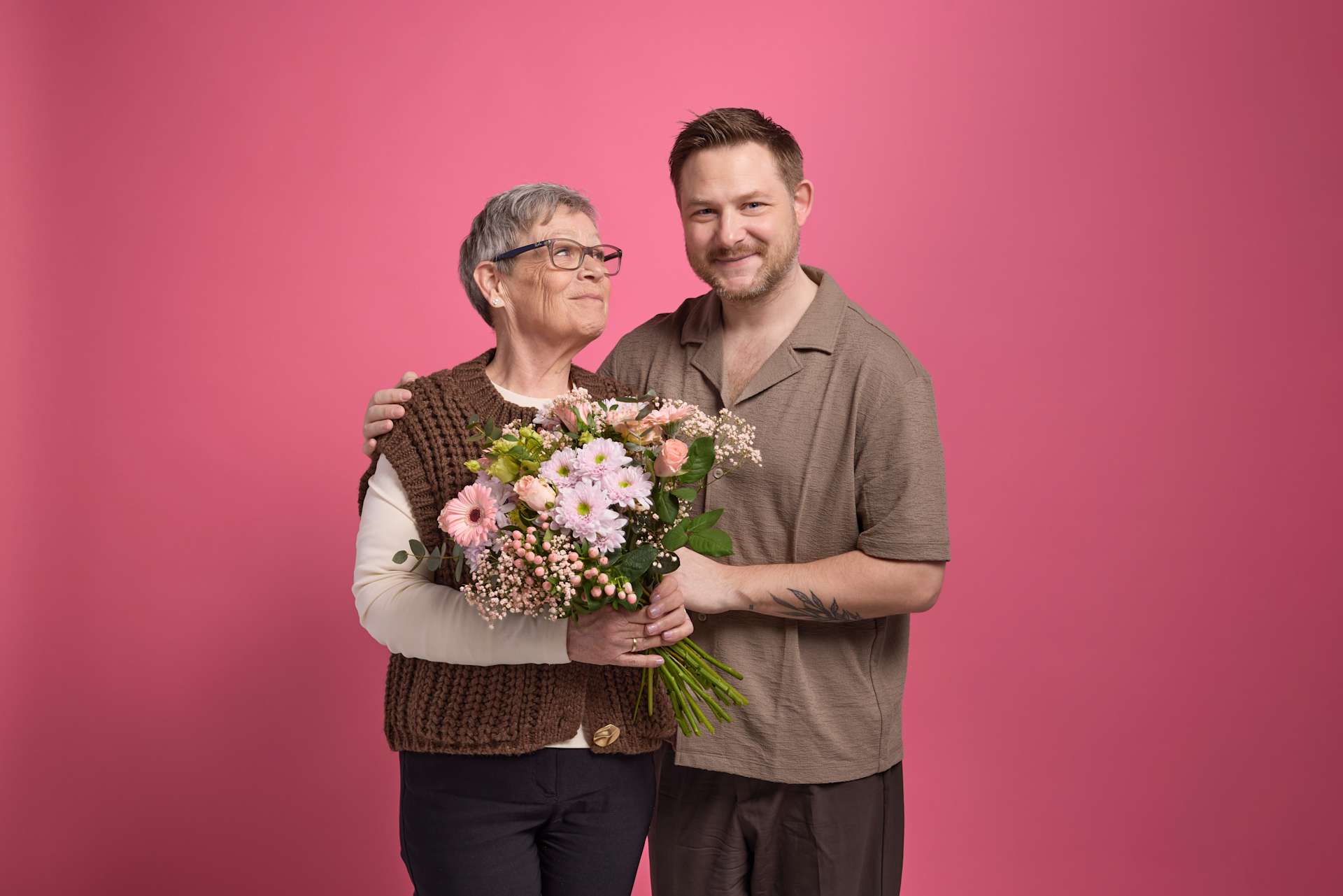 Das Bild zeigt eine ältere Frau und einen jüngeren Mann, die zusammen vor einem leuchtend rosa Hintergrund stehen, wobei die Frau einen Strauß bunter Blumen hält.
