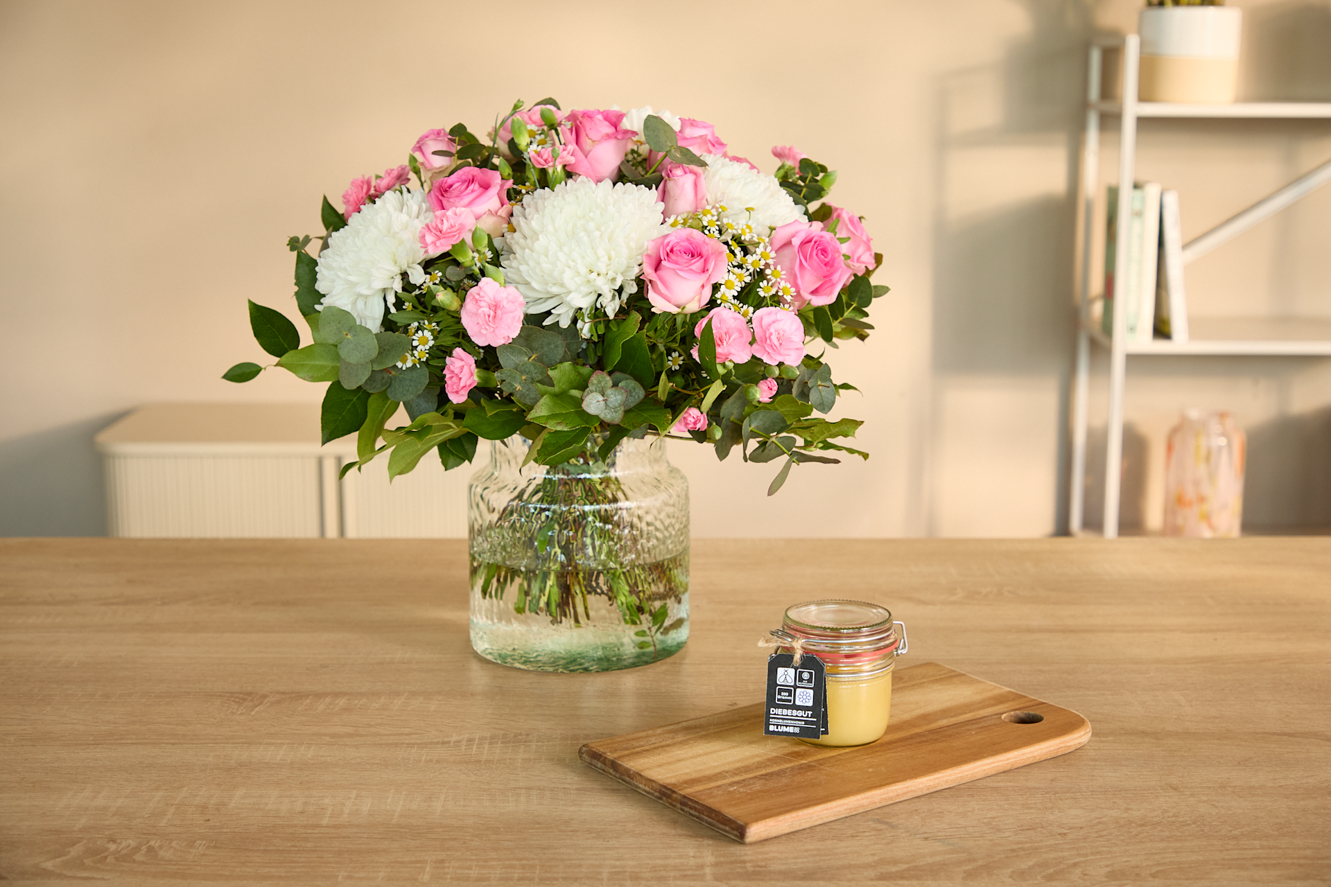 Ein Strauß leuchtend rosa und weißer Blumen in einer Glasvase steht auf einem Holztisch, im Hintergrund sind ein Schneidebrett und Gewürze zu sehen.
