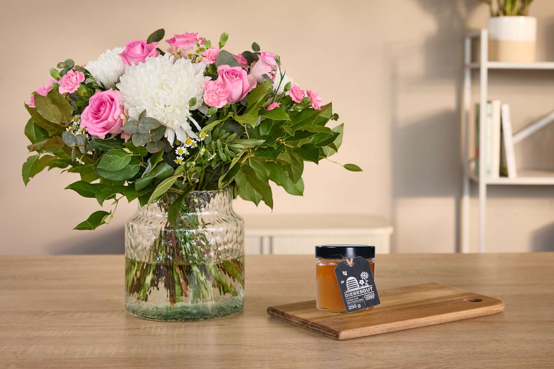 Ein Strauß leuchtend rosa Rosen und weißer Chrysanthemen in einer Glasvase steht auf einem Holztisch, daneben ein kleines Glas Honig und ein Holzbrett.