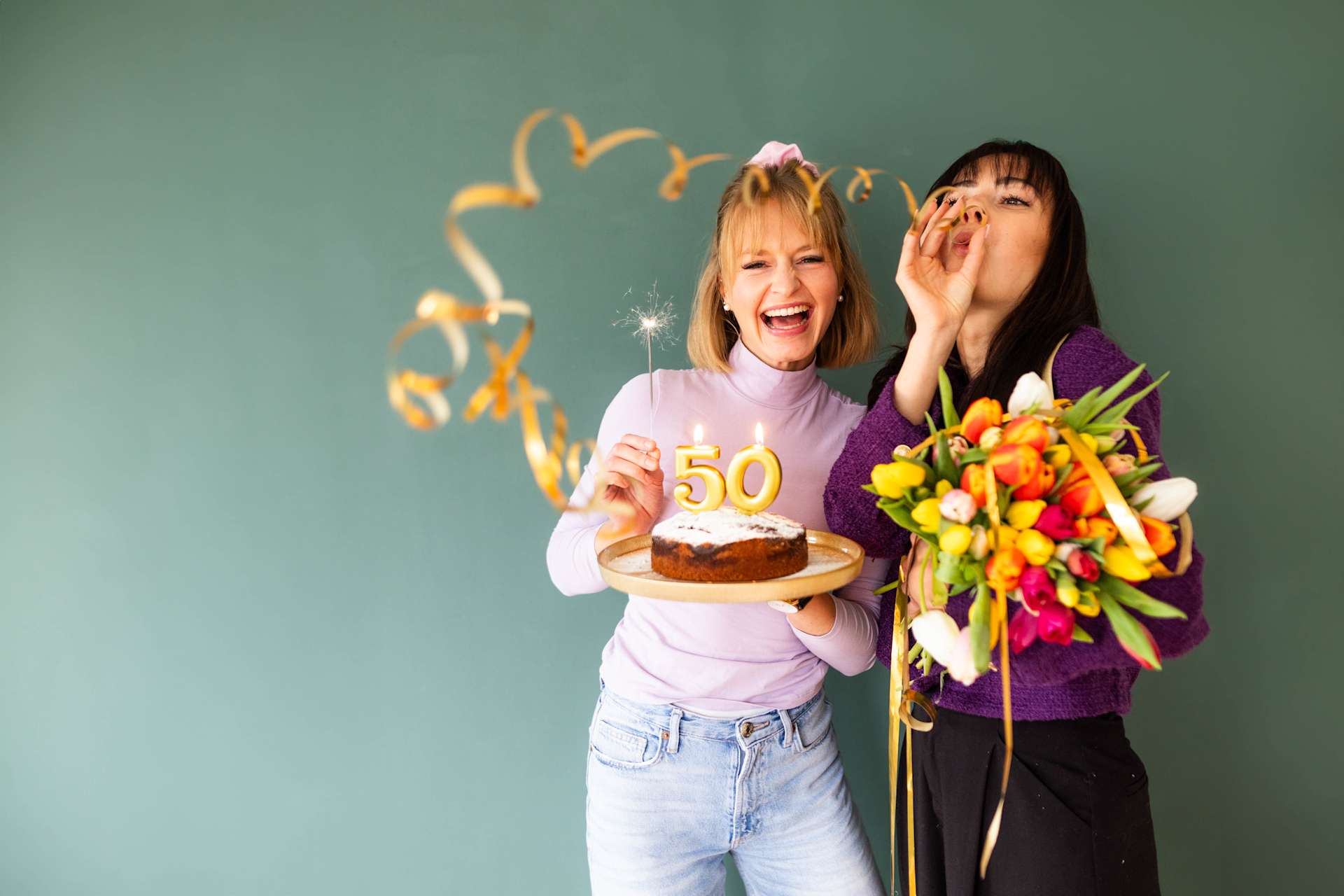 Zwei junge Frauen, von denen die eine eine Geburtstagstorte mit einer „50“-Kerze und die andere einen Blumenstrauß hält, scheinen in einer farbenfrohen Umgebung mit grünem Hintergrund einen runden Geburtstag zu feiern.