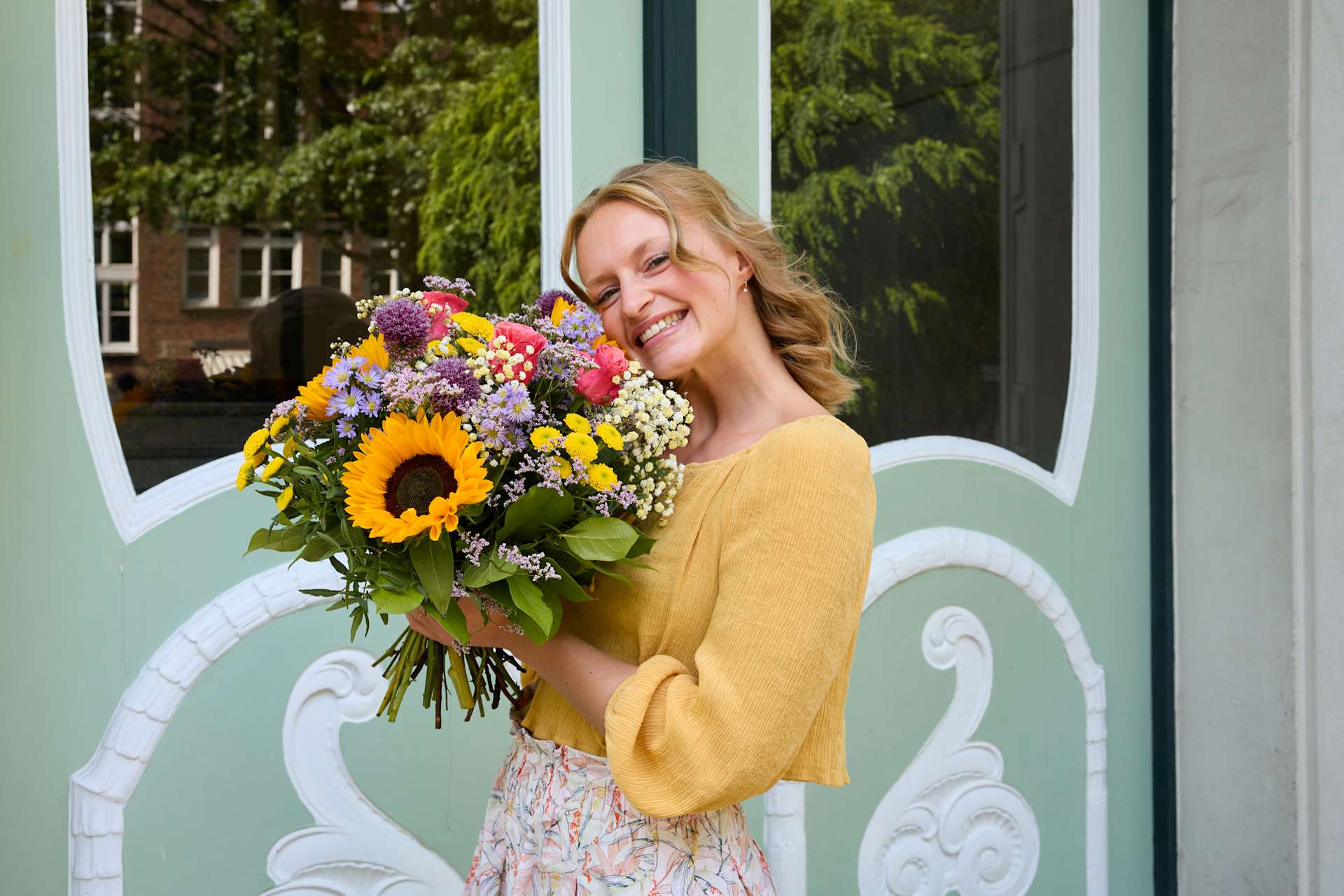 Eine lächelnde Frau in einem gelben Pullover hält einen leuchtenden Blumenstrauß vor einem bunt gemusterten Hintergrund.