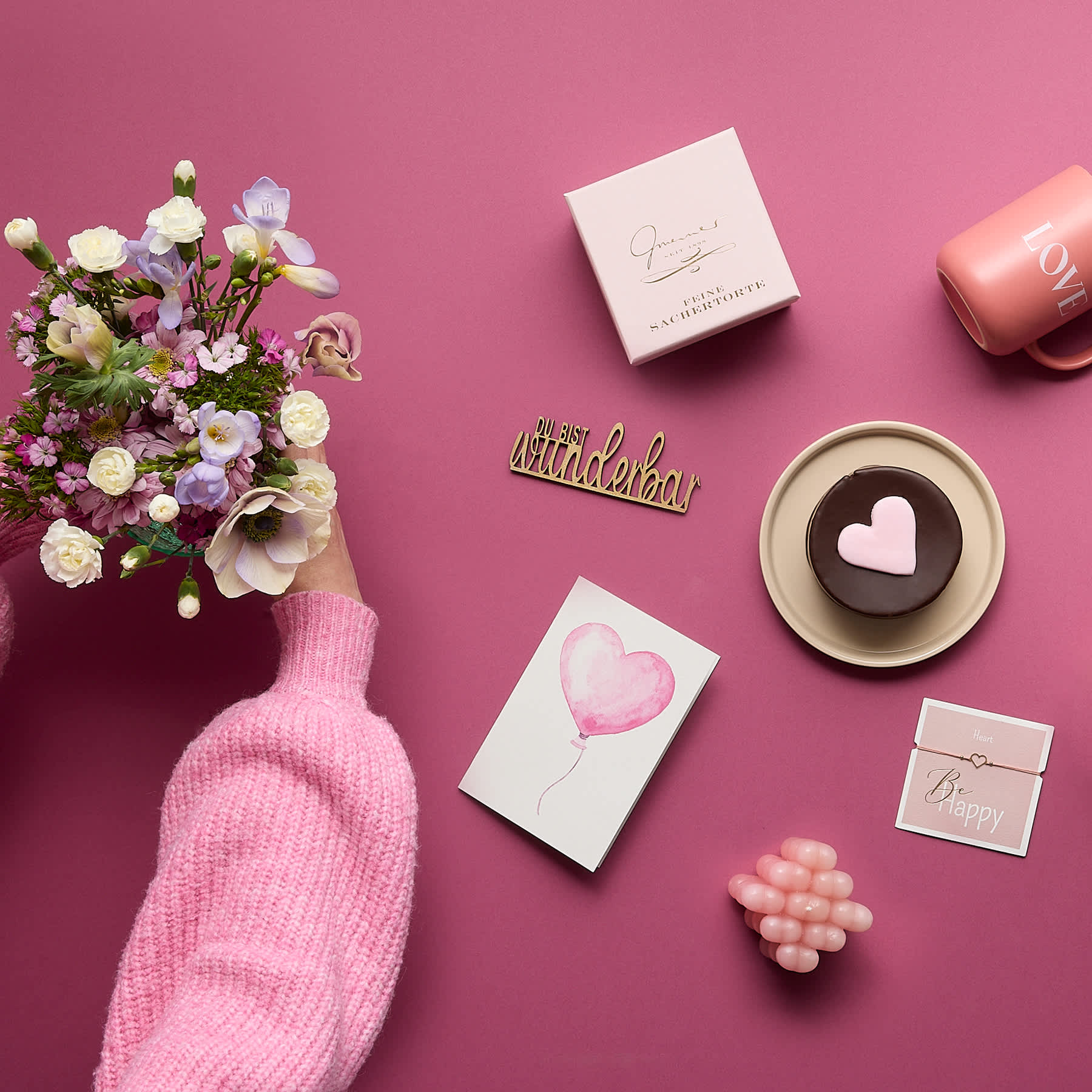 Ein Strauß bunter Blumen, ein rosa Pullover, eine herzförmige Schokolade und verschiedene andere romantische Gegenstände sind auf einem leuchtend rosa Hintergrund arrangiert.