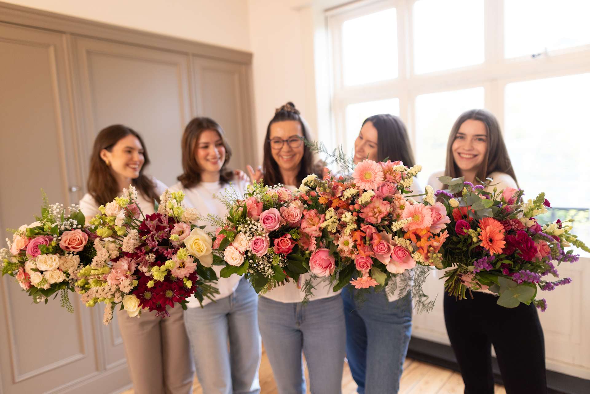 Eine Gruppe von fünf jungen Frauen hält große, leuchtende Sträuße bunter Blumen in einem hellen, luftigen Raum mit großen Fenstern.