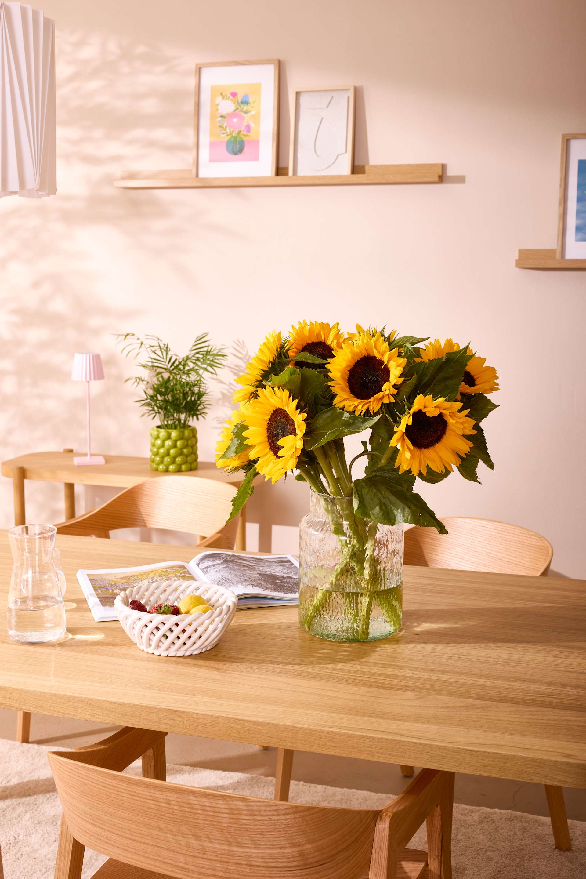 Ein gemütlicher Essbereich mit einem Holztisch und Stühlen, geschmückt mit einem farbenfrohen Strauß Sonnenblumen in einer Glasvase und gerahmten Kunstwerken auf den Regalen im Hintergrund.