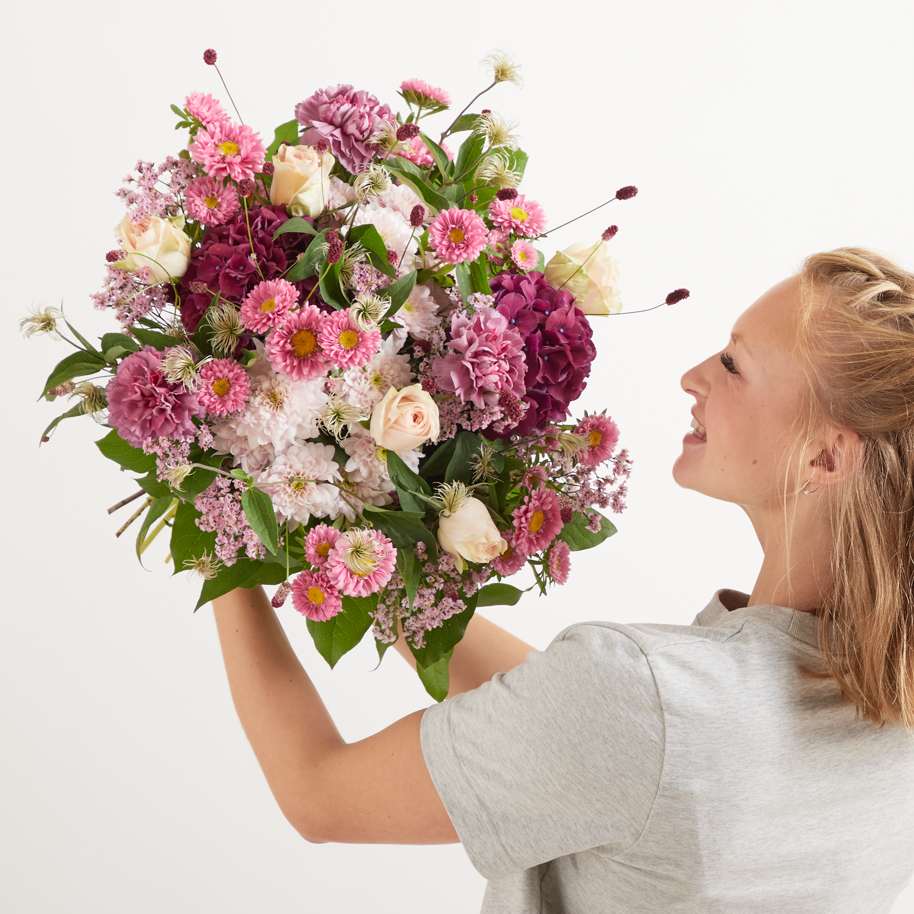 Eine Frau hält vor einem schlichten weißen Hintergrund einen farbenfrohen Strauß rosa und weißer Blumen, darunter Pfingstrosen, Rosen und Chrysanthemen.