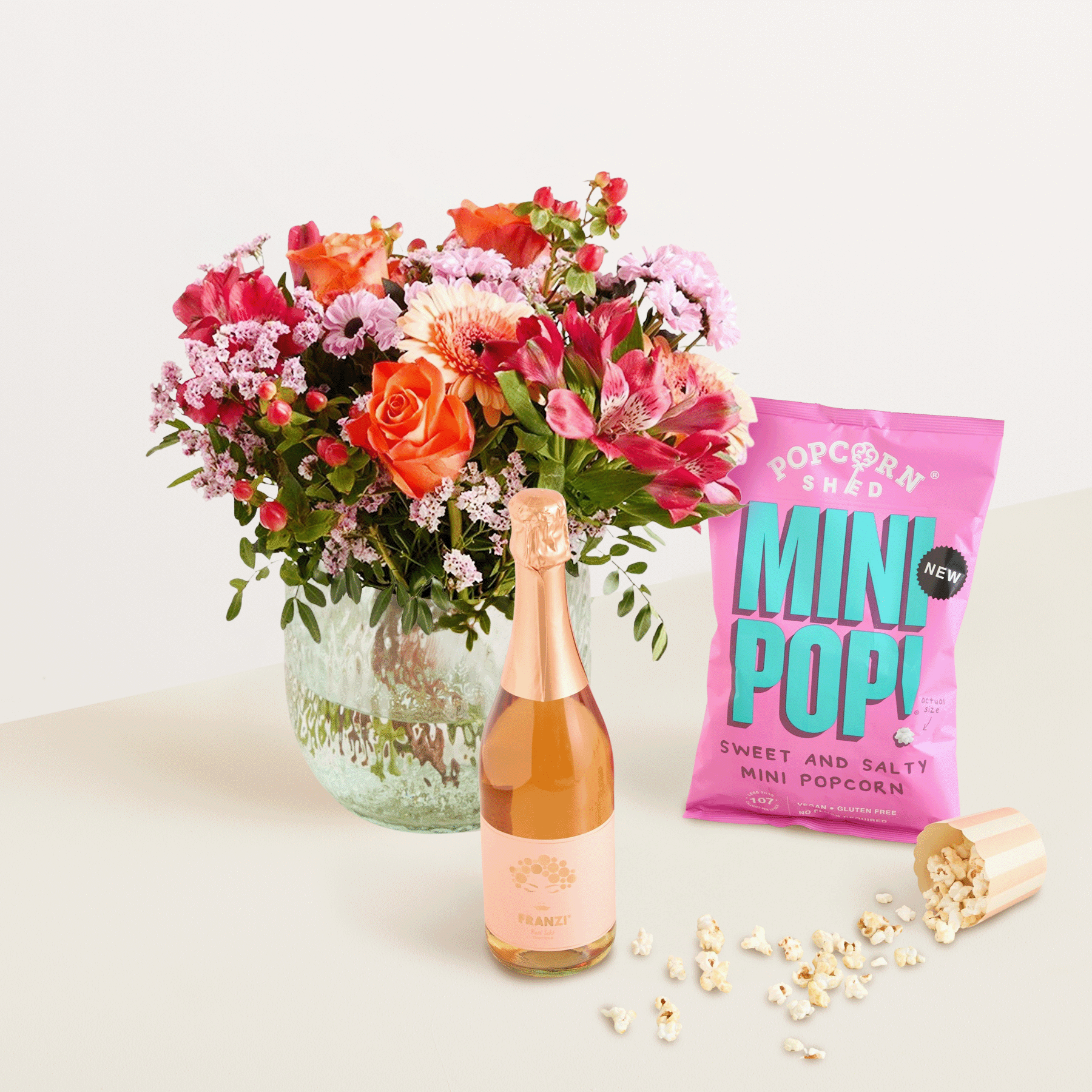 Ein farbenfroher Strauß bunter Blumen, darunter Rosen und Nelken, steht neben einer Flasche Sekt und einer Tüte Mini-Popcorn auf einem schlichten Hintergrund.